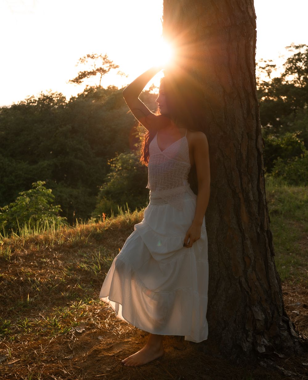 Woman in white dress leaning against tree trunk with stunning golden sunset rays behind her.