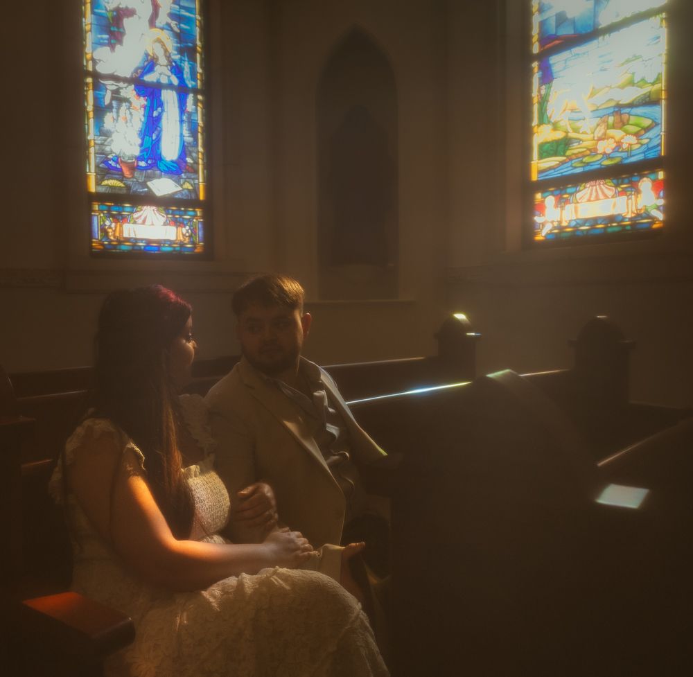 Bride and groom sitting in church pews with colorful stained glass windows illuminating the scene.