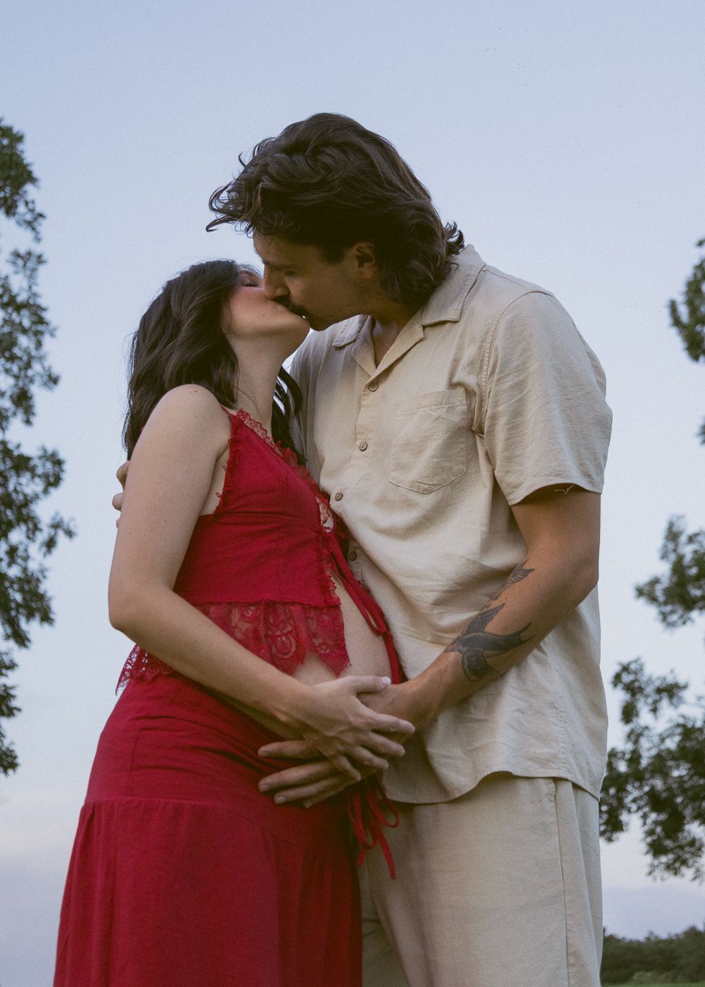 Couple sharing a kiss outdoors, woman in red dress with visible baby bump, man in beige shirt holding her waist.