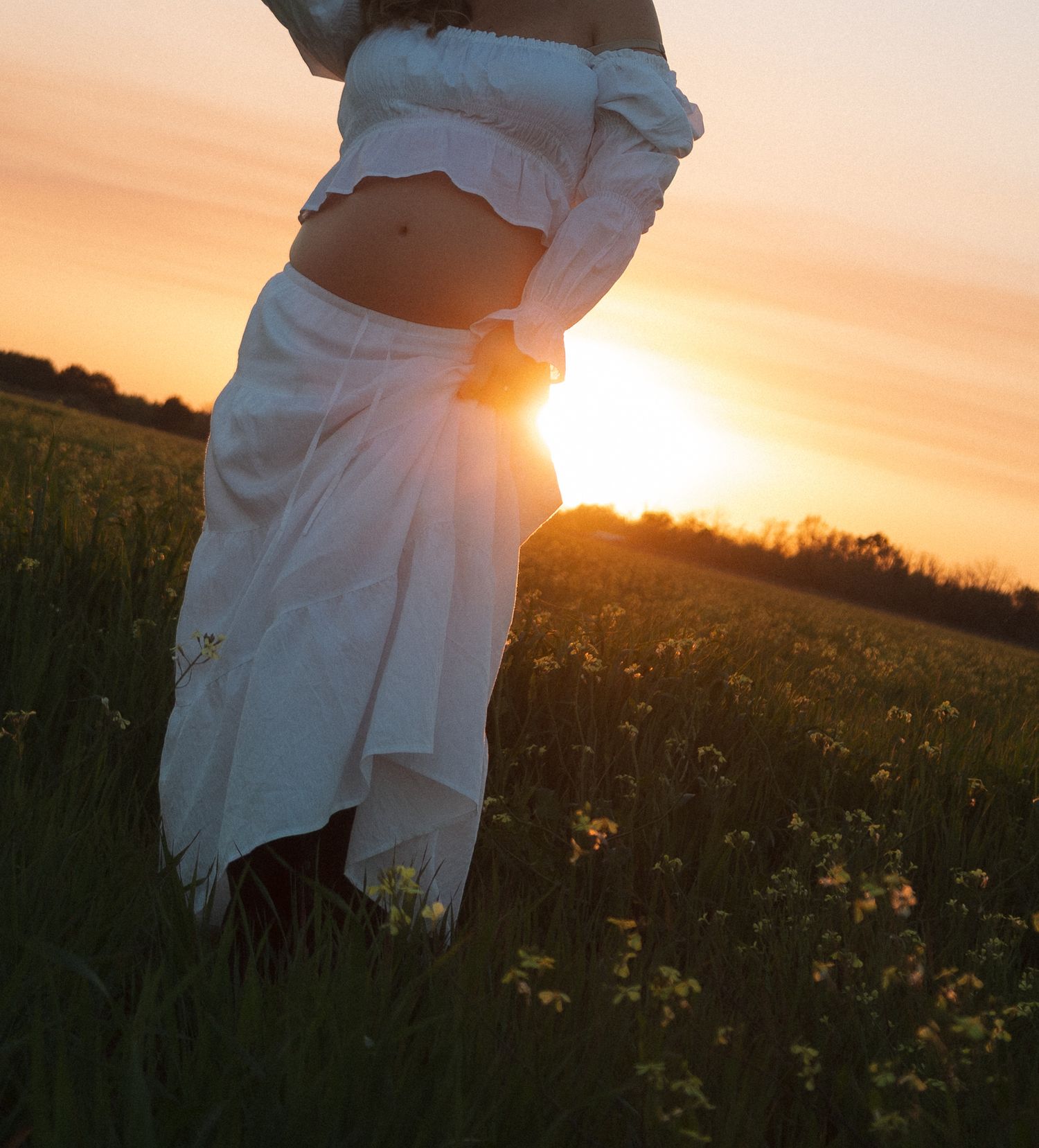 Pregnant woman in white outfit backlit by vibrant sunset in flower field.