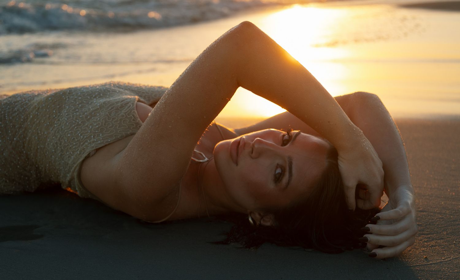 Woman lying on beach sand smiling with hands in hair during golden sunset.