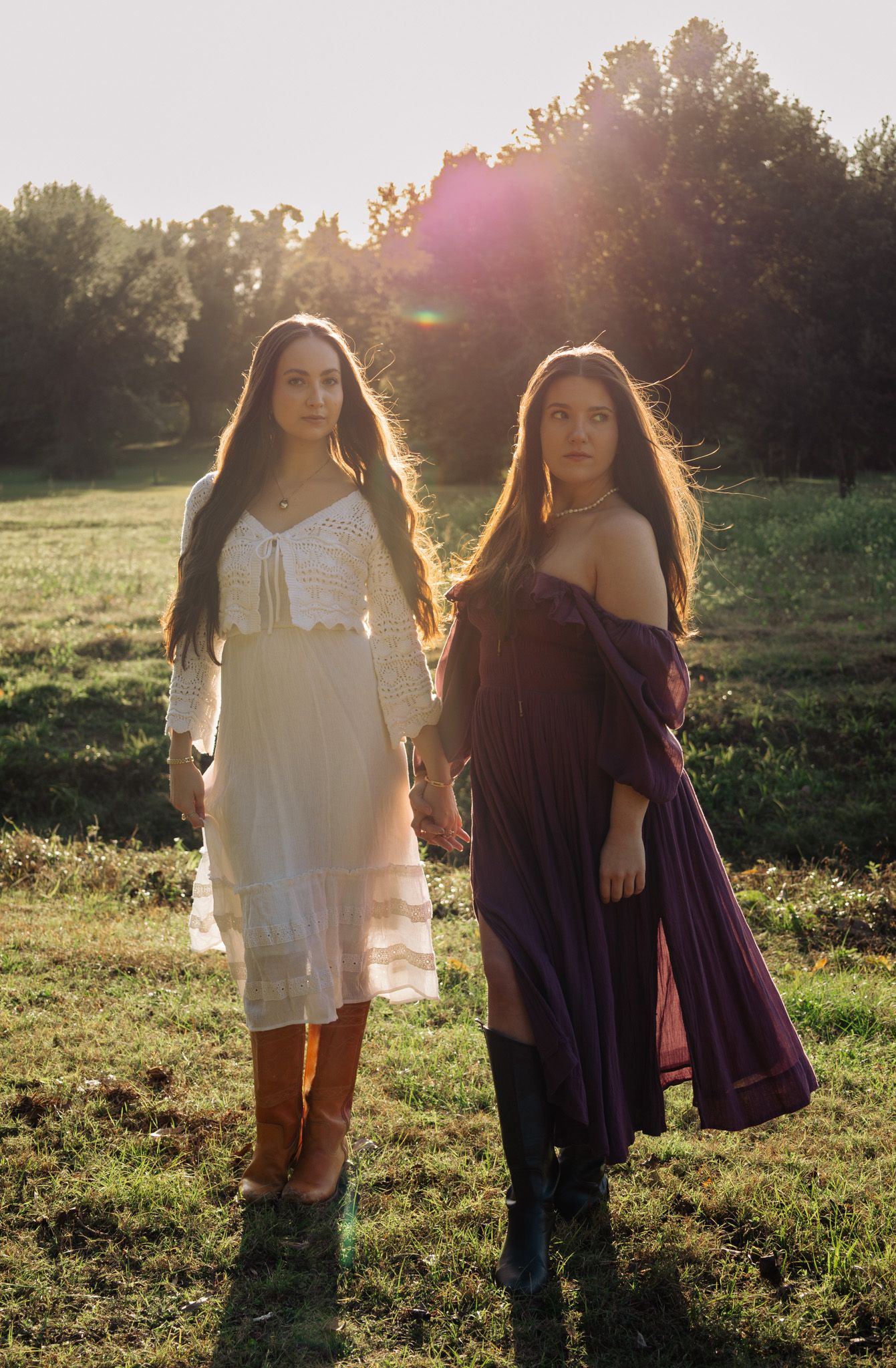 Two young women in boho dresses holding hands in a sunlit field with golden hour lens flare.