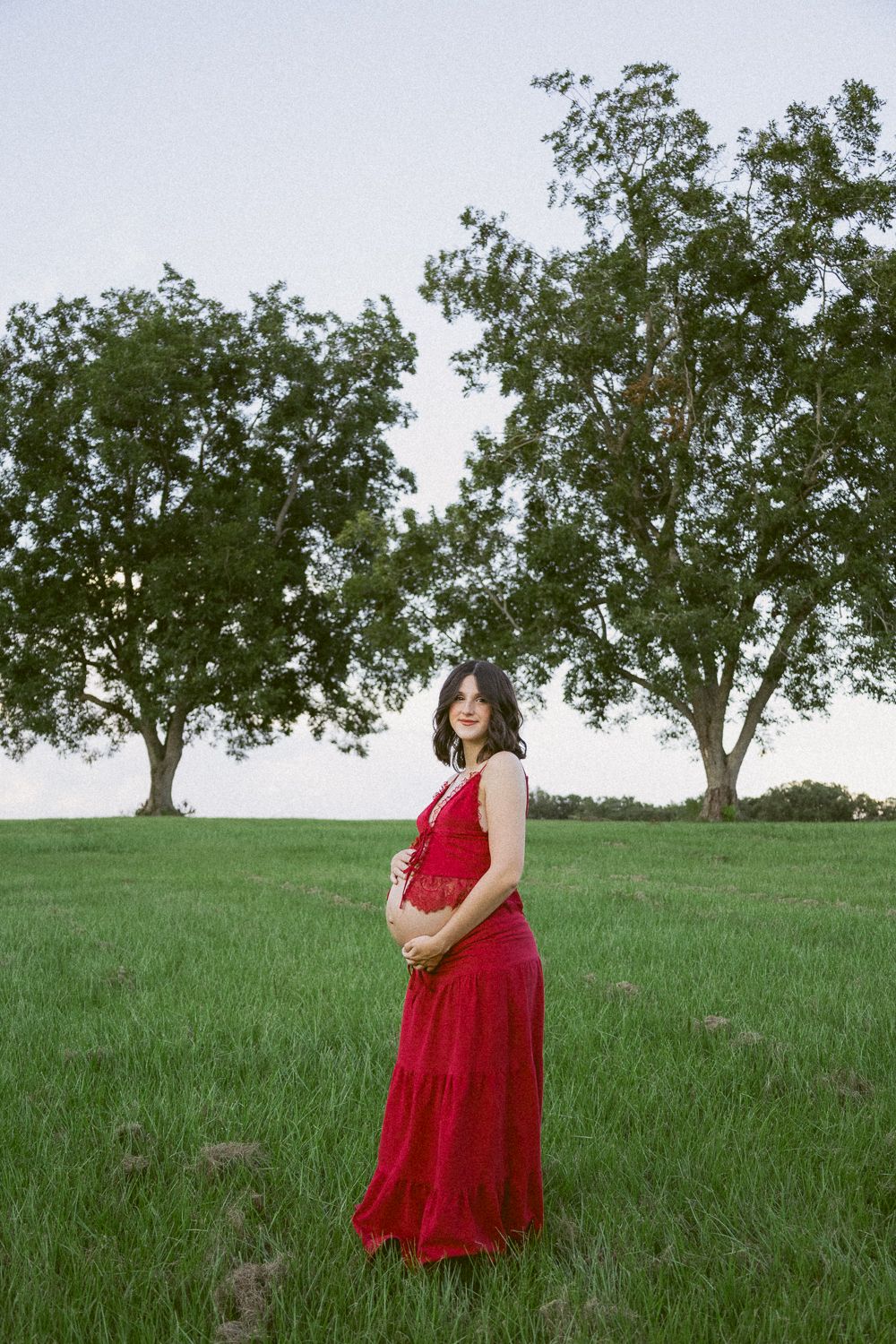 Pregnant woman in elegant red lace maxi dress standing in lush green field with trees during maternity shoot.