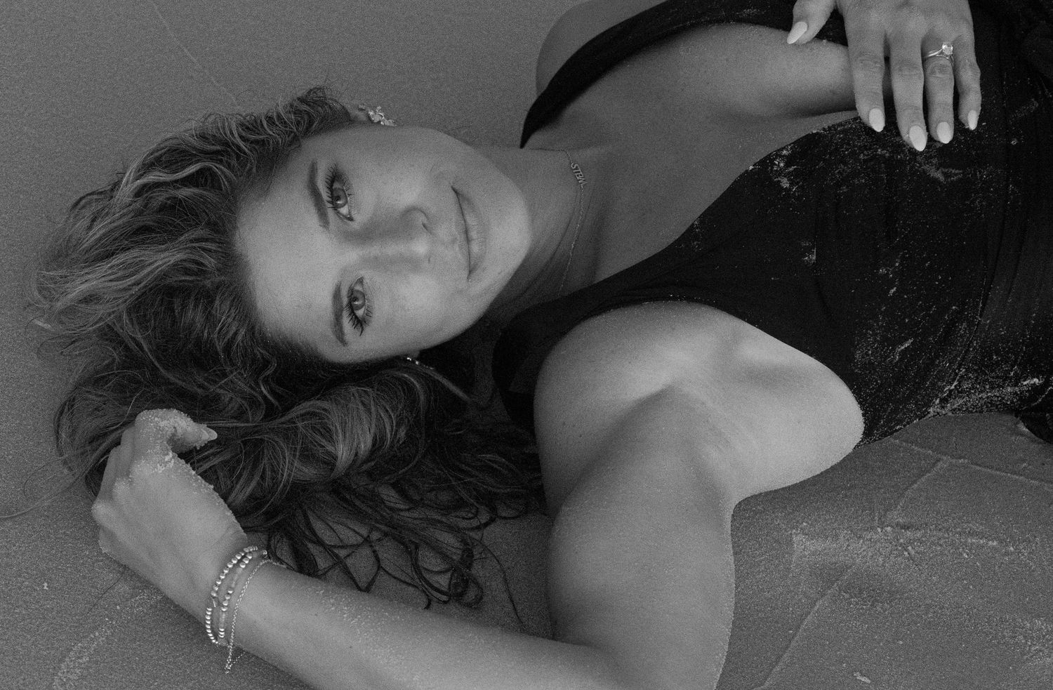 Black and white photo of a woman lying on sand, smiling with wavy hair and bracelets.