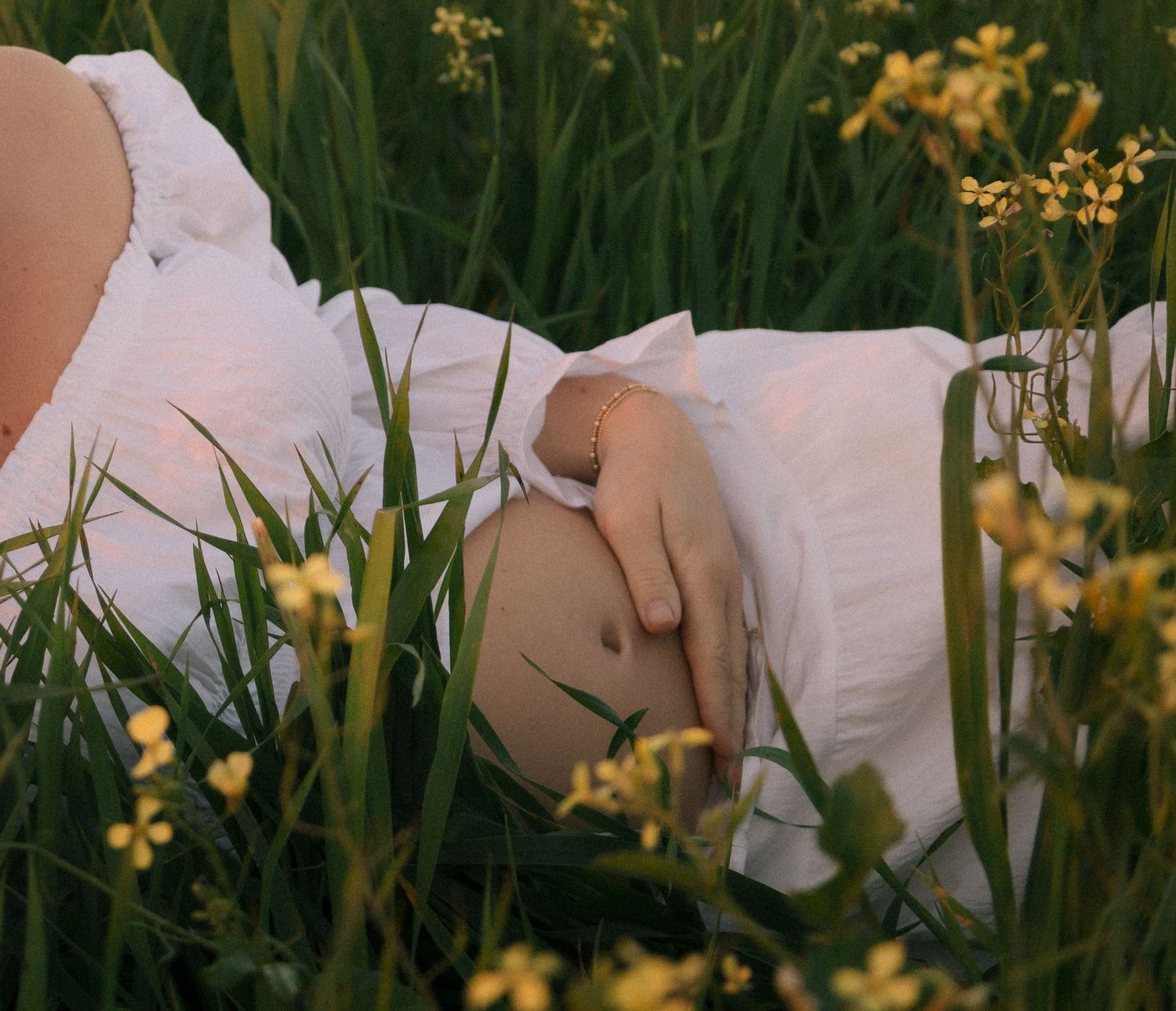Pregnant woman lying in green grass field with yellow wildflowers, hands on belly.
