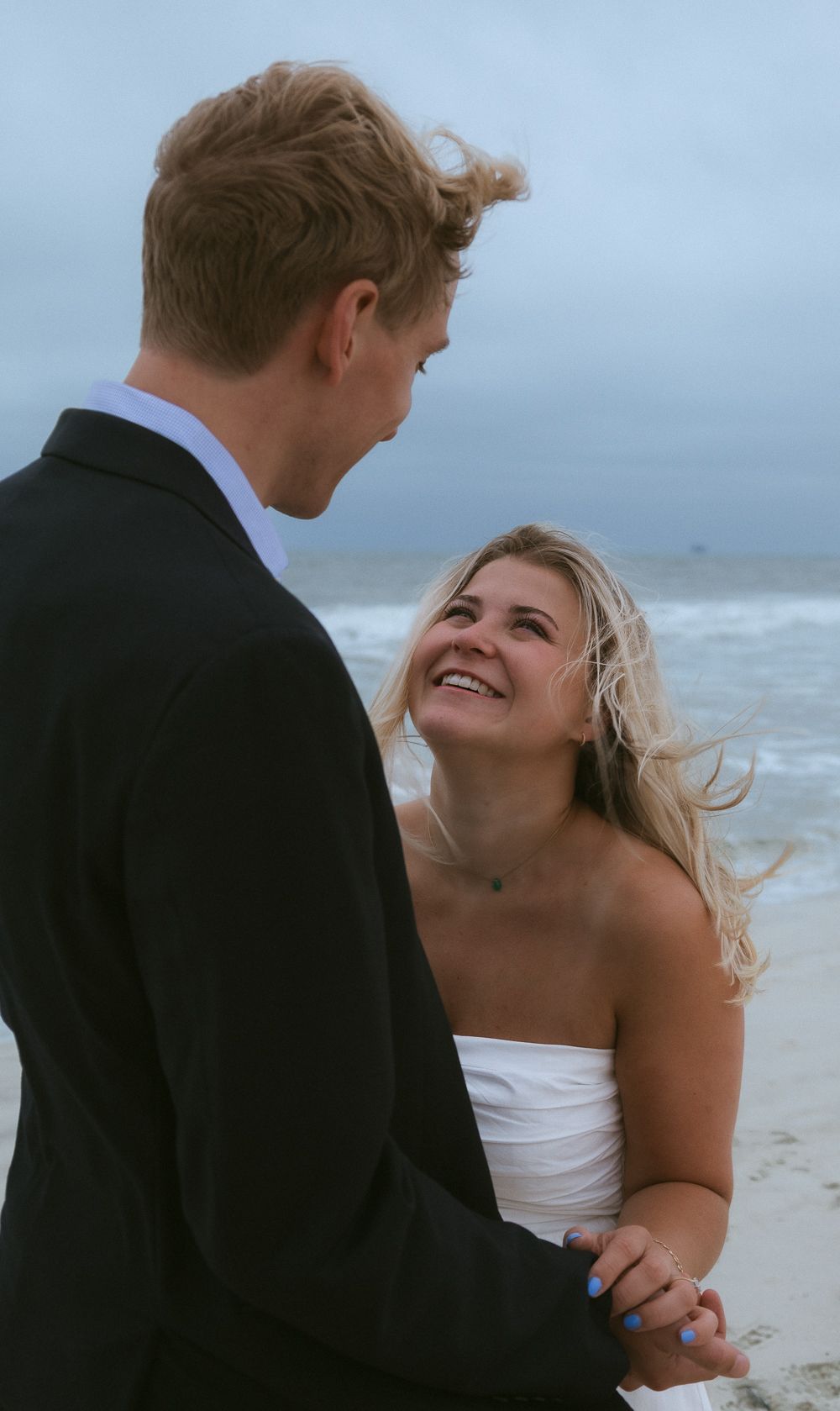 Smiling blonde woman in white dress gazes up at man in black suit on windy beach.
