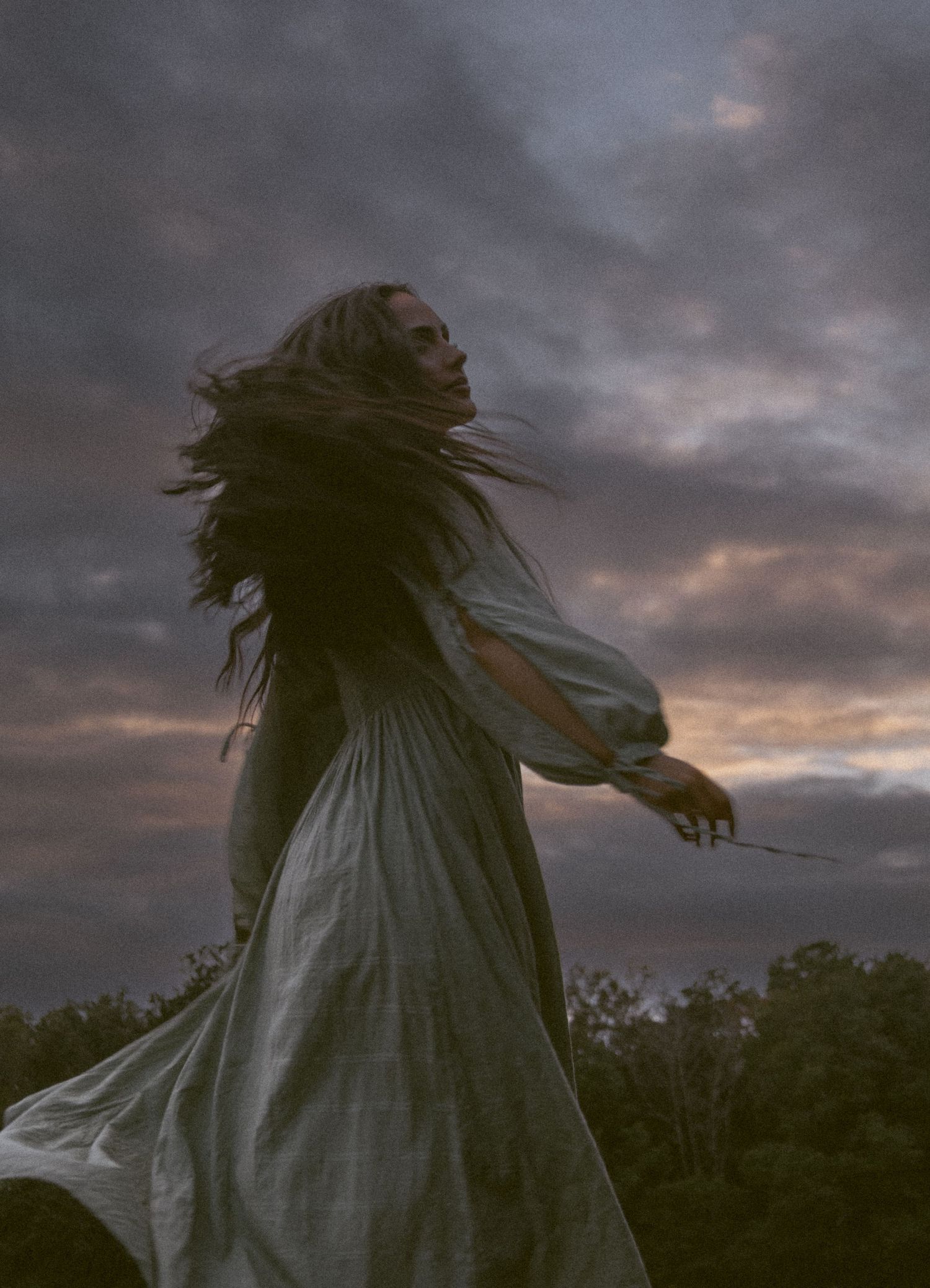 Woman in flowing white dress with hair blowing dramatically against stormy sunset sky outdoors.