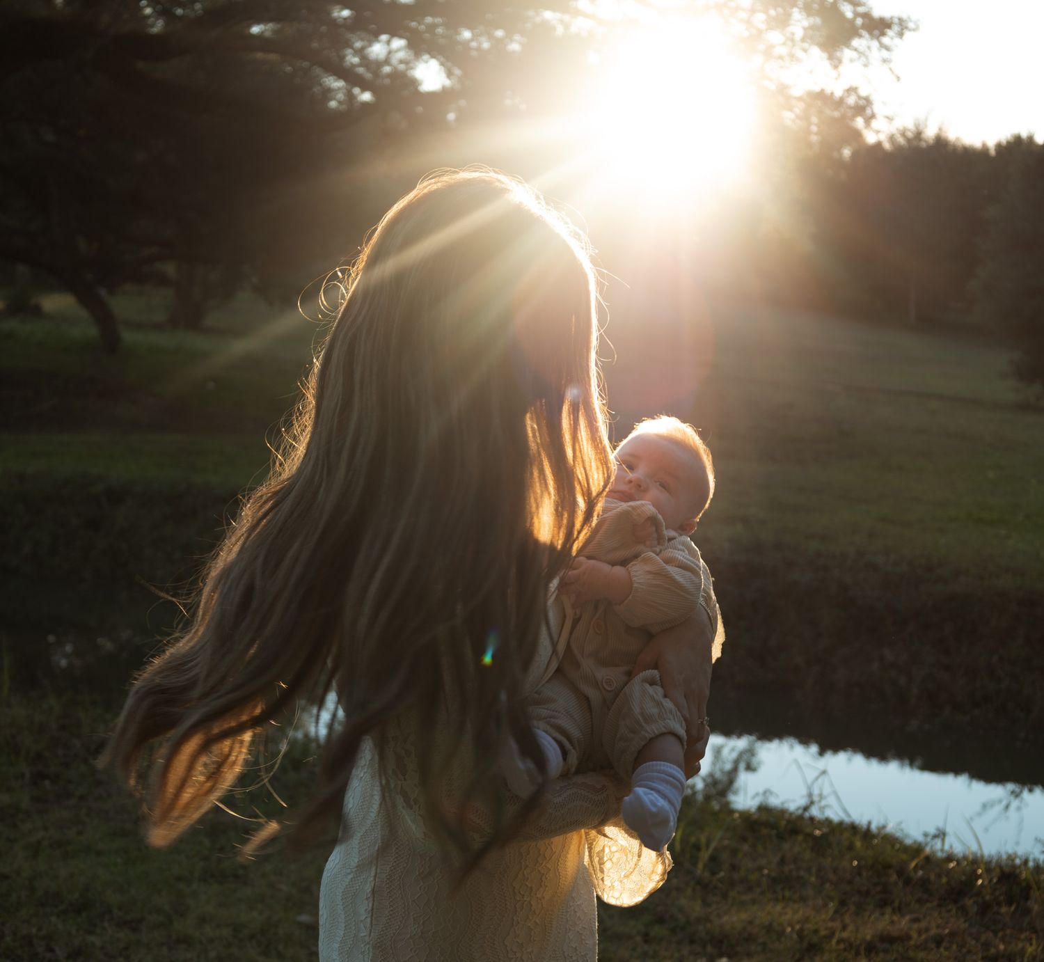 Mother holding baby while standing in glowing golden hour sunlight near pond.