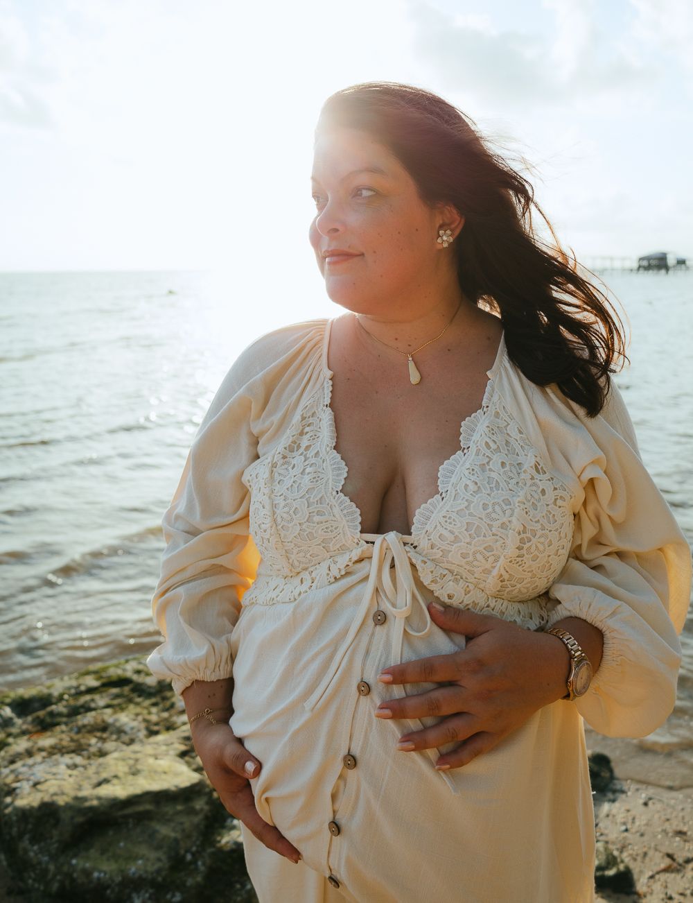 Pregnant woman in cream lace dress cradling baby bump on rocky beach at golden hour with ocean backdrop.