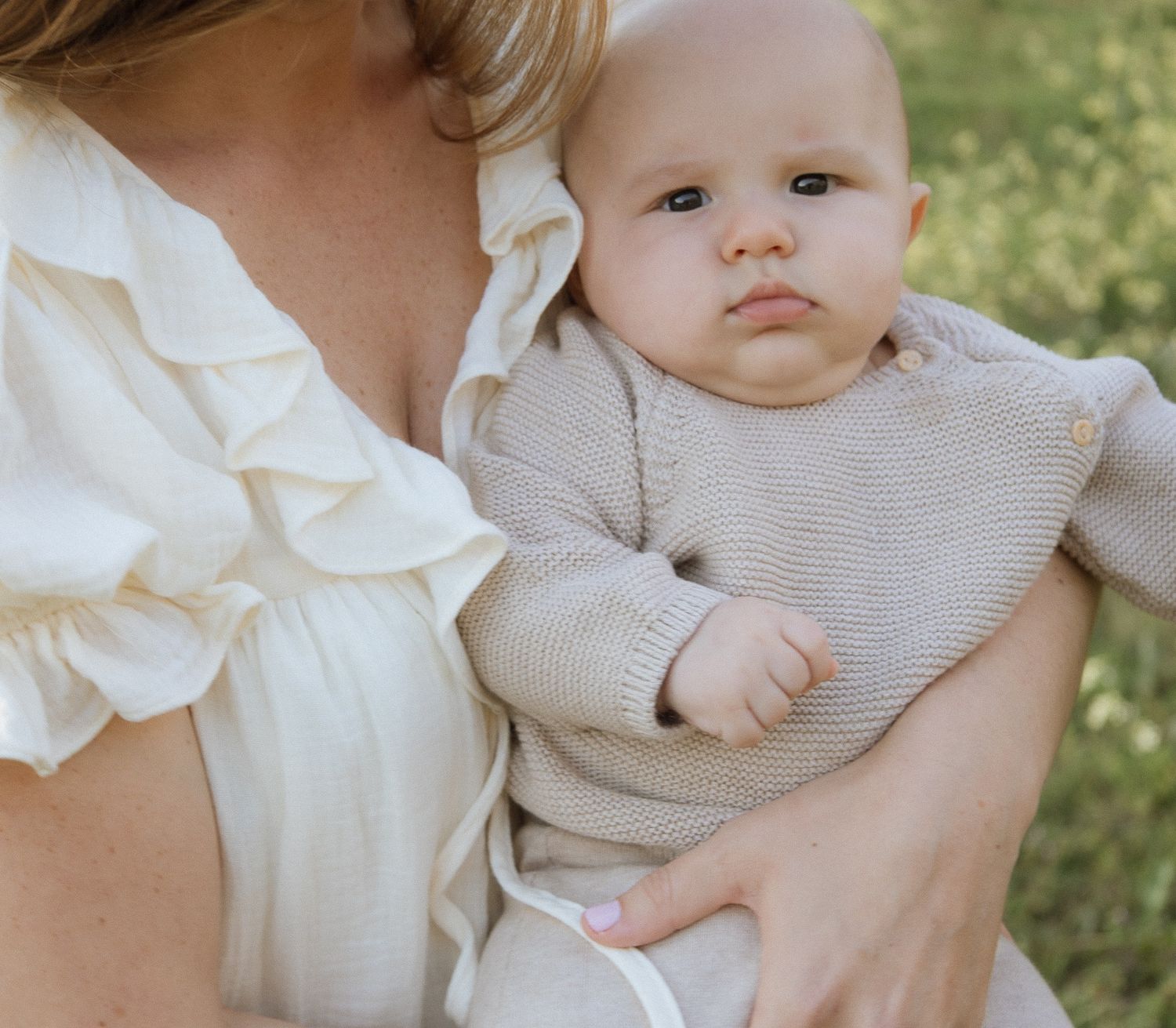 Close-up of alert newborn baby in beige knit romper being held by mother in white ruffled dress.