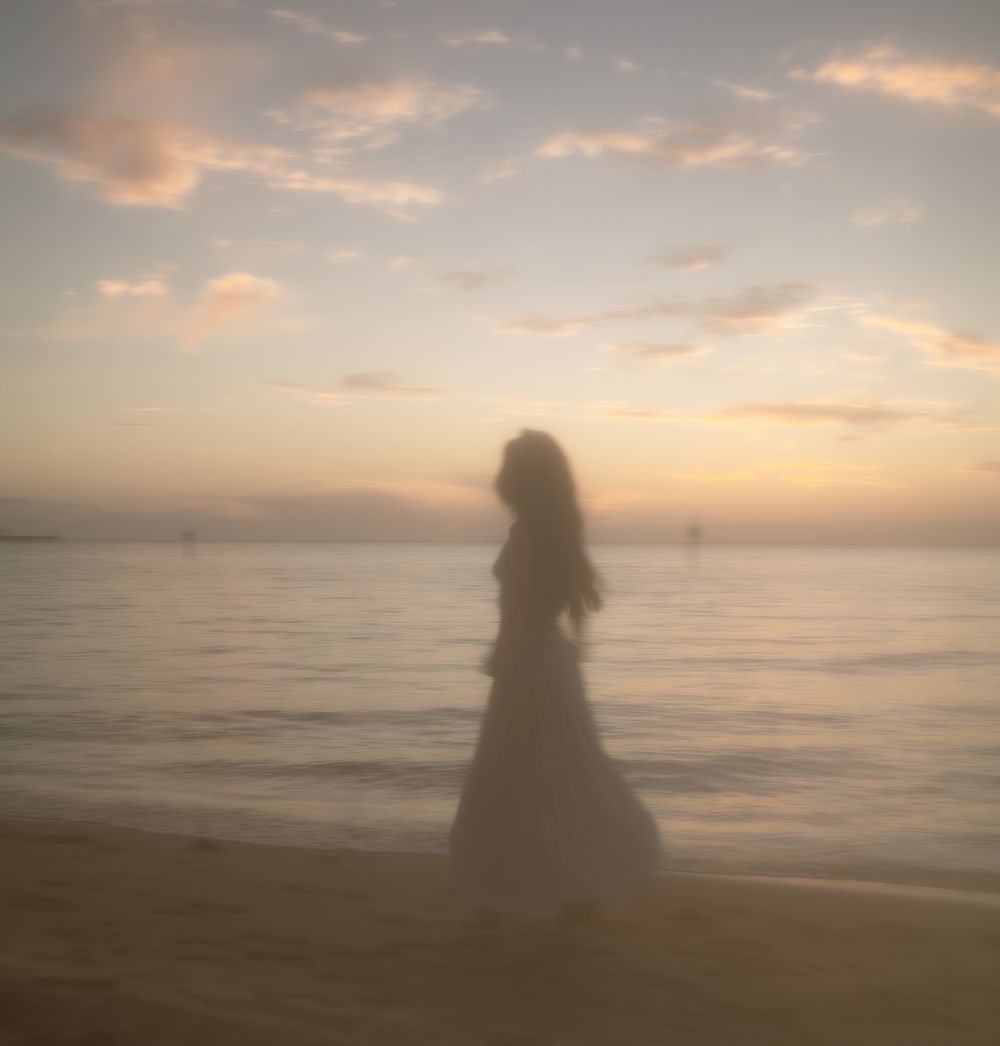 Silhouette of woman in dress standing on beach at golden sunset with calm ocean backdrop.