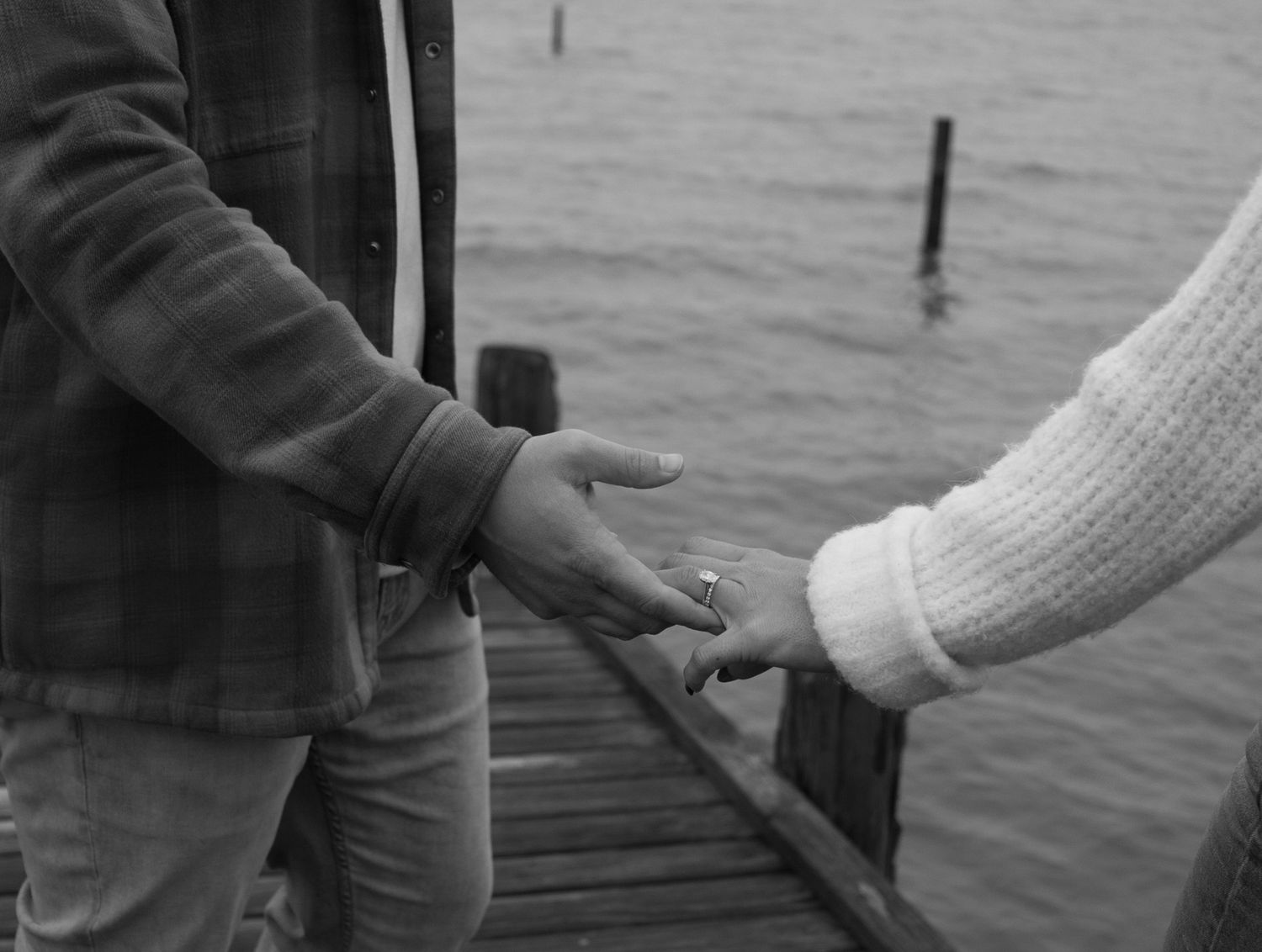 Black and white close-up of couple holding hands on a wooden pier near water.