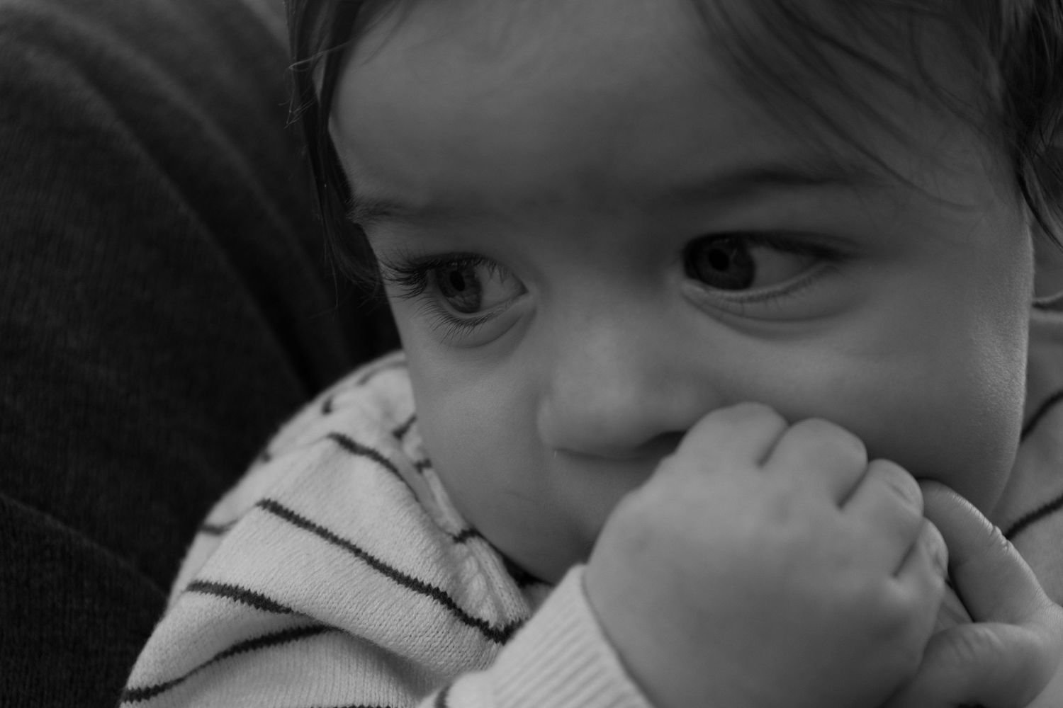 Black and white close-up of a young toddler with dark eyes chewing on their hand.