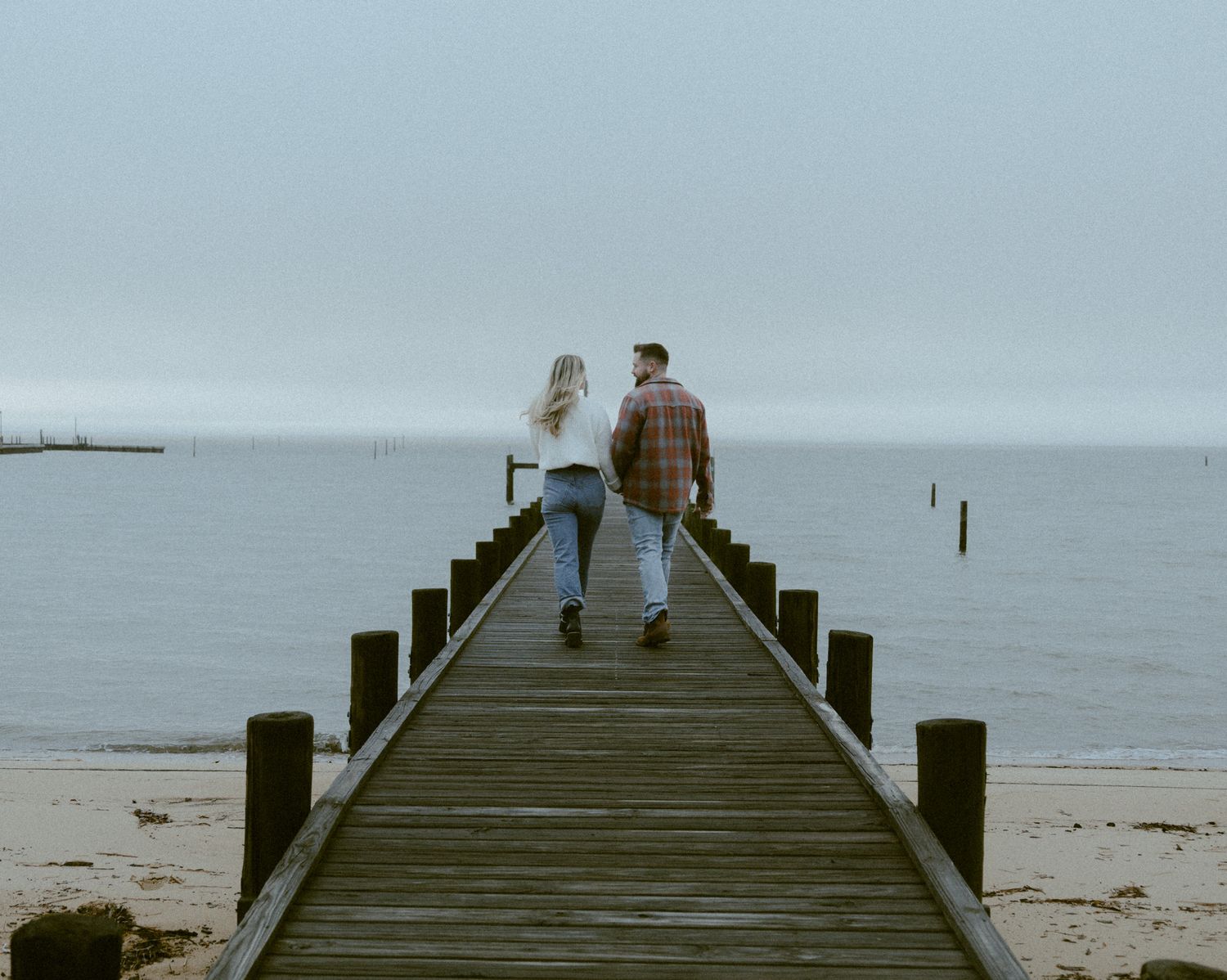 Couple walking hand in hand down a wooden pier toward calm ocean water under overcast sky.