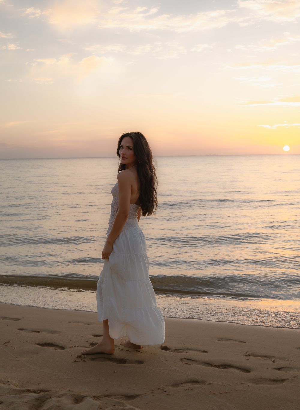 Elegant woman in flowing white maxi dress walking barefoot on beach leaving footprints at sunset.