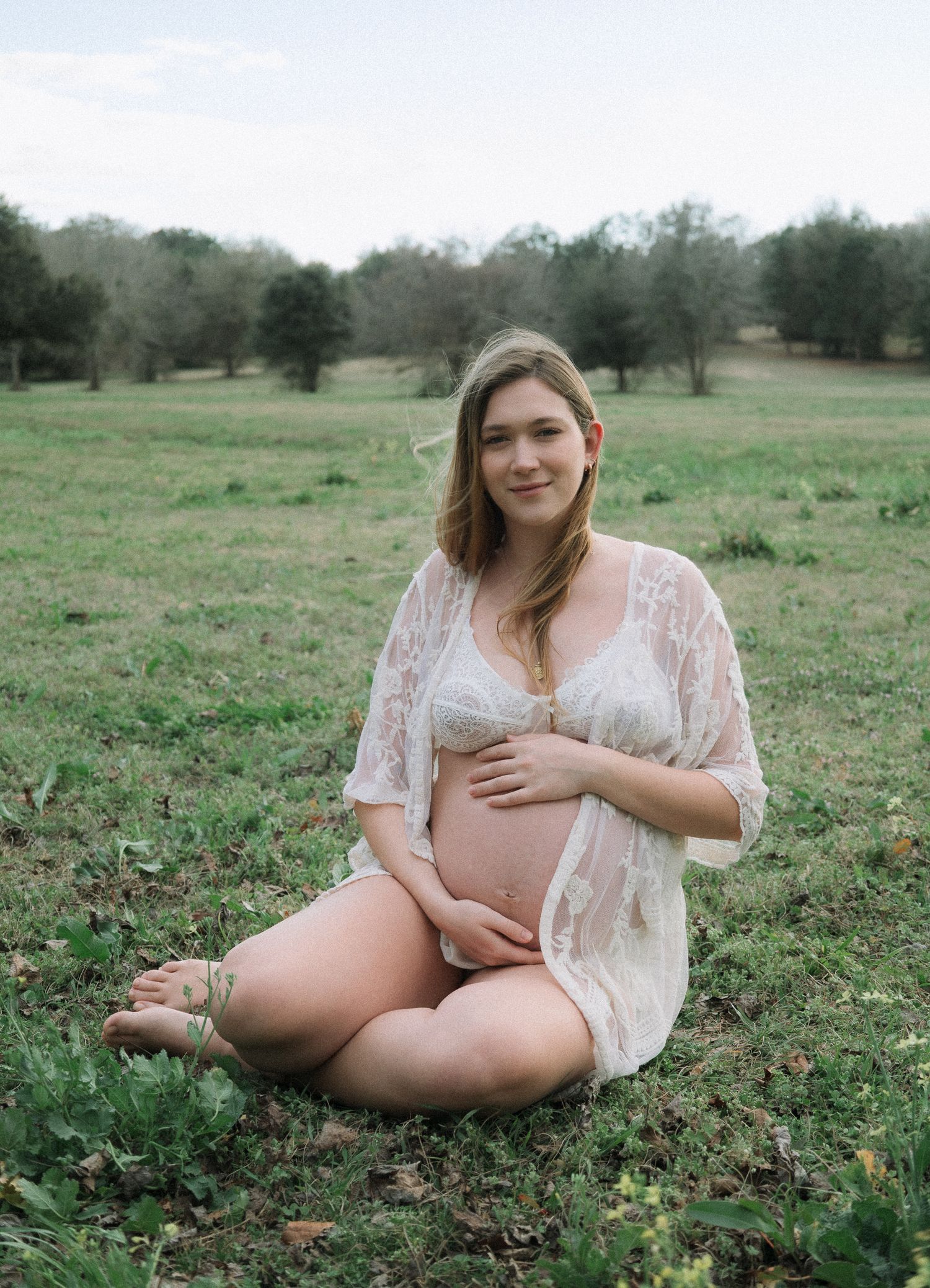 Pregnant woman in lace robe and white bra sitting in green field during maternity photoshoot.