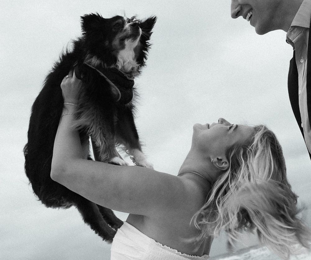 Black and white photo of woman lifting small dog toward man outdoors.