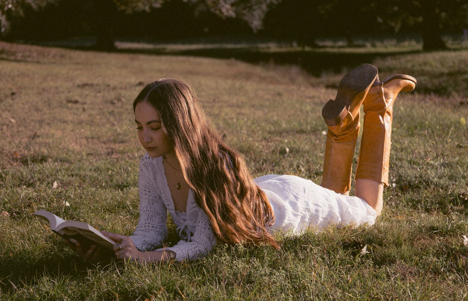 Young woman in white dress and yellow boots lying on grass reading a book in golden sunlight.