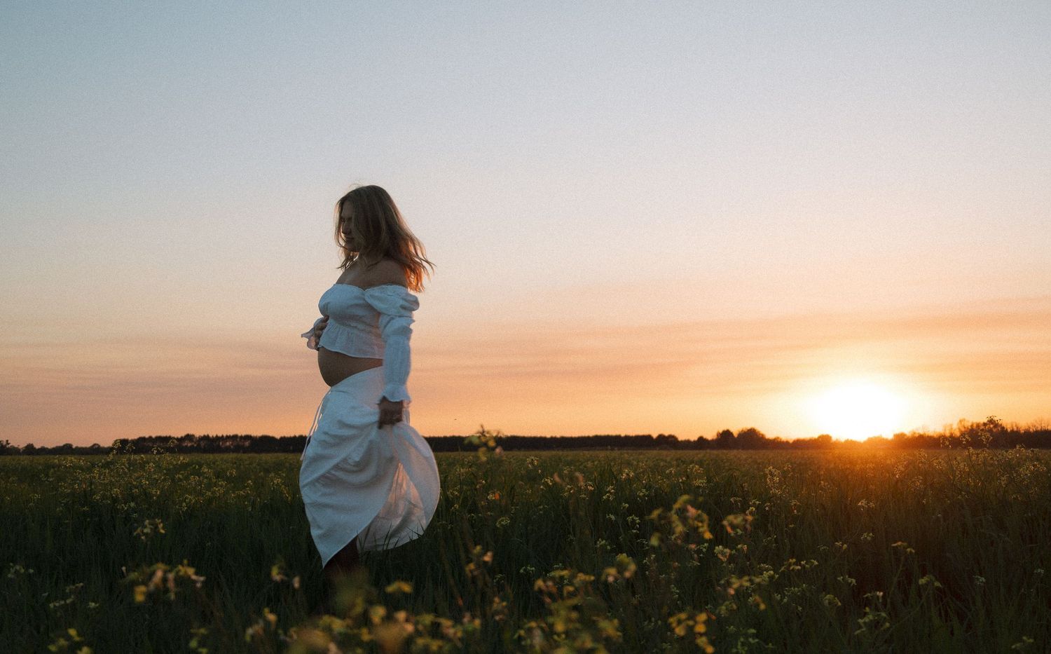 Pregnant woman in white dress standing in blooming field during golden hour sunset.