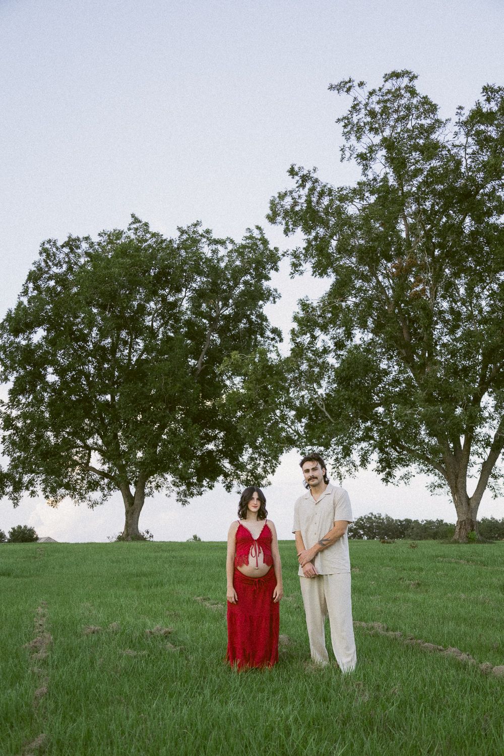 Pregnant couple standing in green field with large trees, woman in red maternity dress, man in cream outfit.
