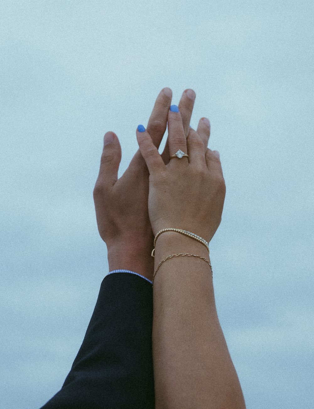 Two hands raised against a blue sky, one wearing a diamond engagement ring and gold bracelets with blue painted nails.