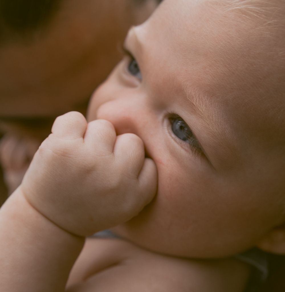 Close-up of an adorable baby with blue eyes sucking on tiny fist, captured in soft warm tones.