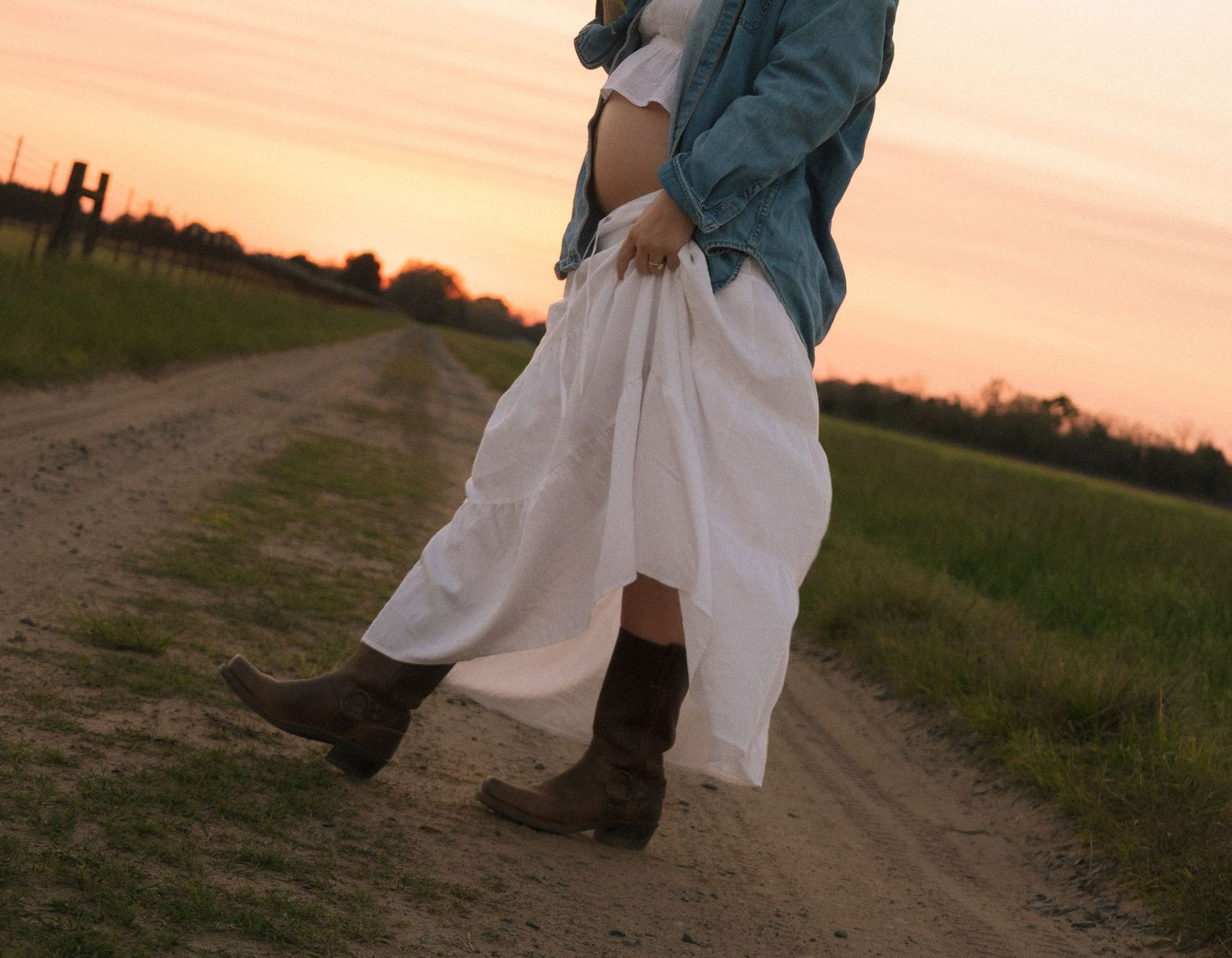 Pregnant woman in white skirt and denim jacket wearing cowboy boots on dirt road.