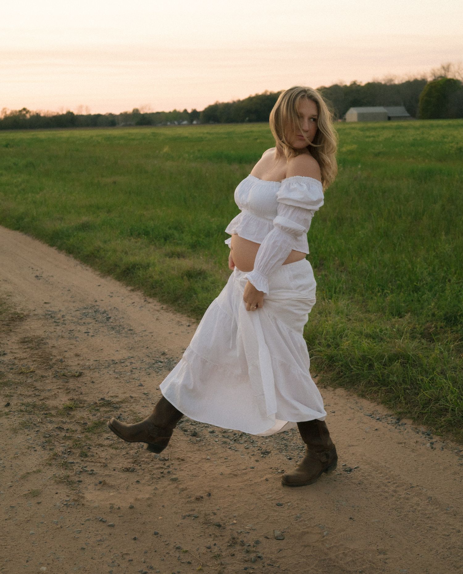 Pregnant woman in white off-shoulder dress and cowboy boots walking on rural dirt road.
