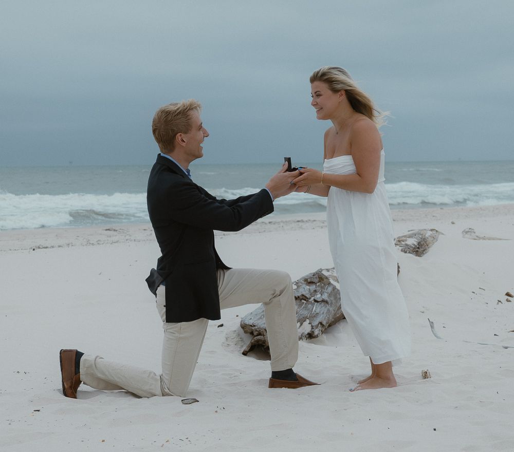 Man on one knee proposing to a woman on a sandy beach with ocean waves in the background.