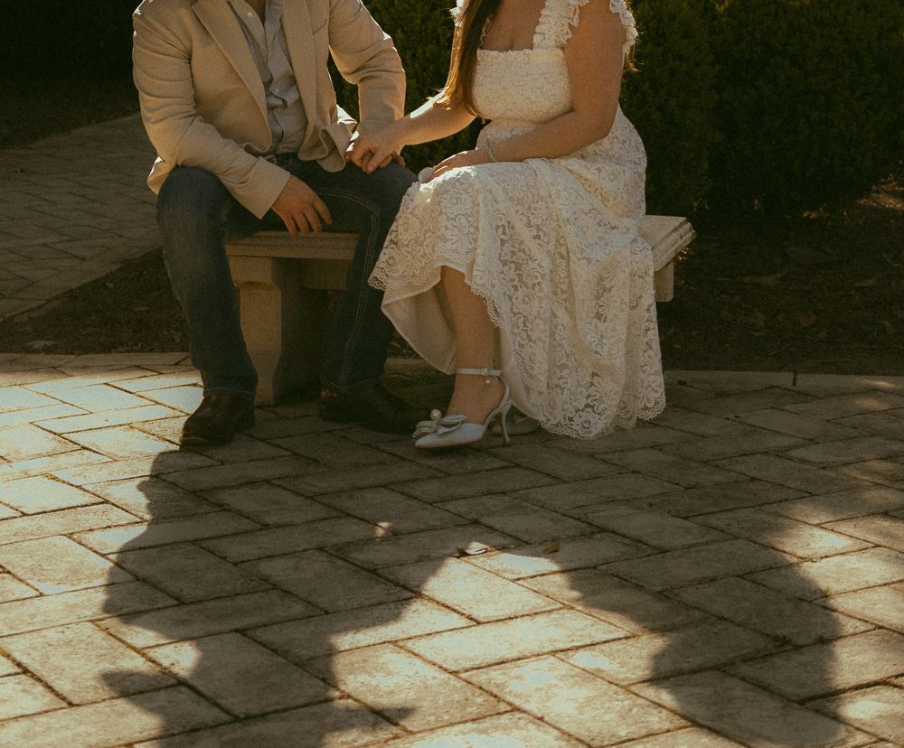 Bride in white lace dress and groom in tan suit holding hands while seated on outdoor bench.