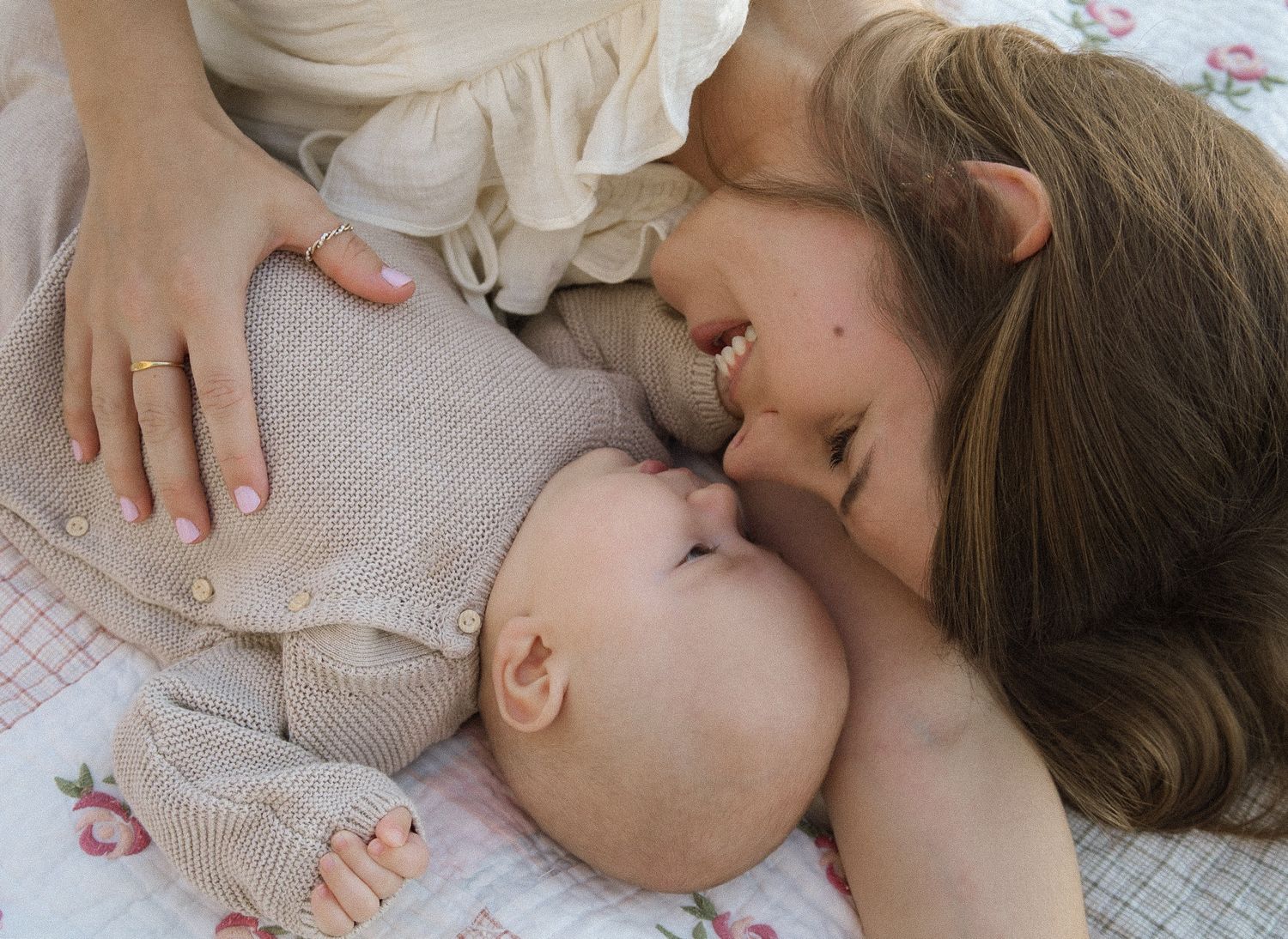 Loving mother in cream dress lying on floral quilt giving gentle kiss to smiling newborn baby.