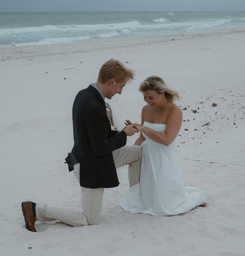 Man kneels on sandy beach placing engagement ring on smiling woman's finger during romantic proposal.