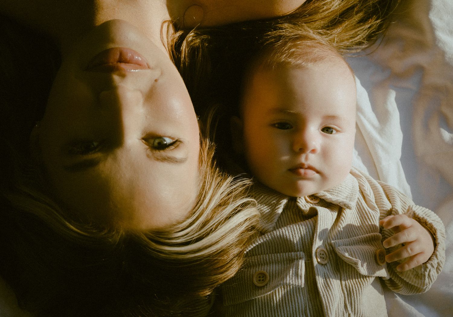 Mother and baby lying together on white bedding, sharing a tender moment in warm light.