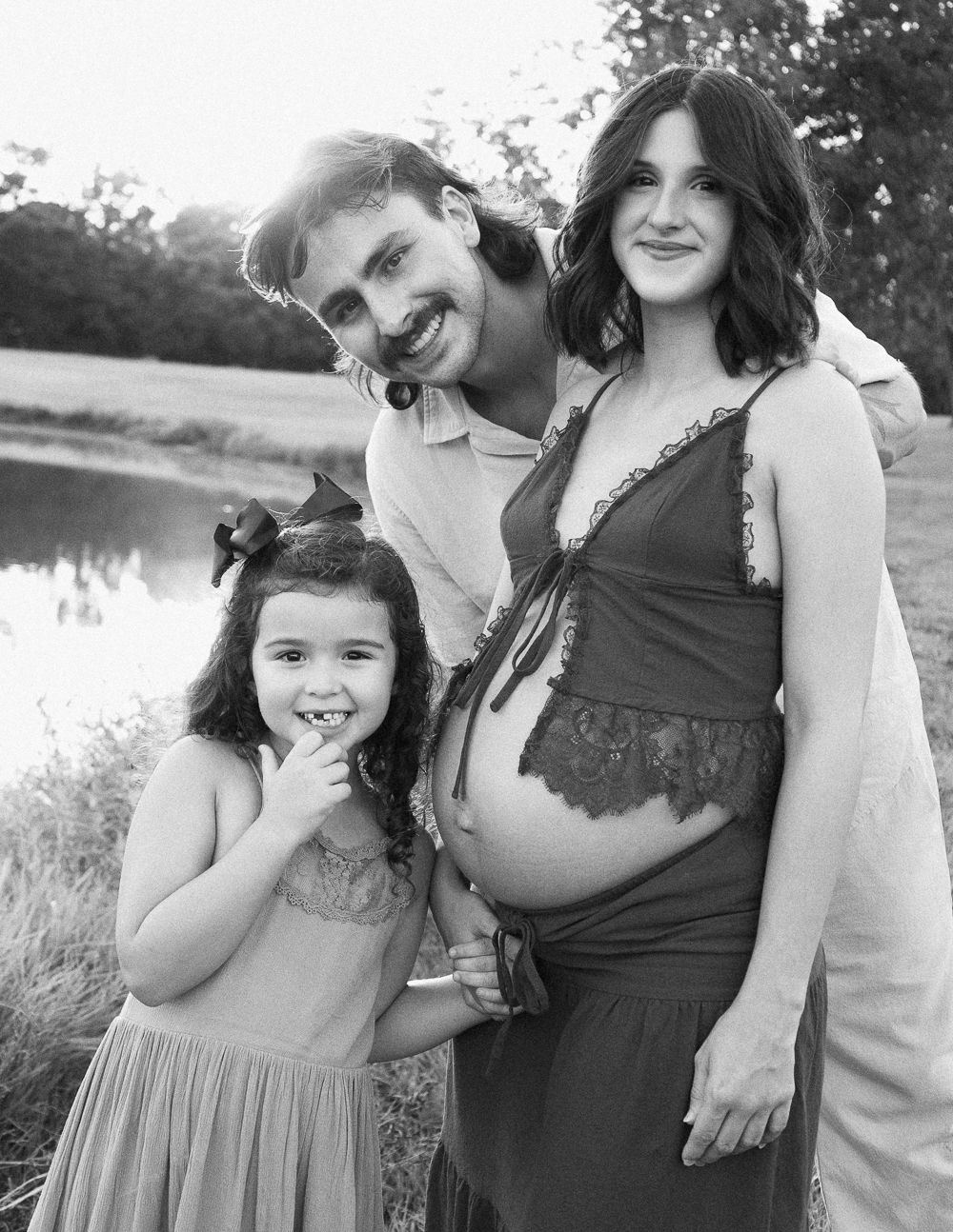 Black and white maternity photo of a smiling family with a young girl and pregnant mother outdoors near a lake.