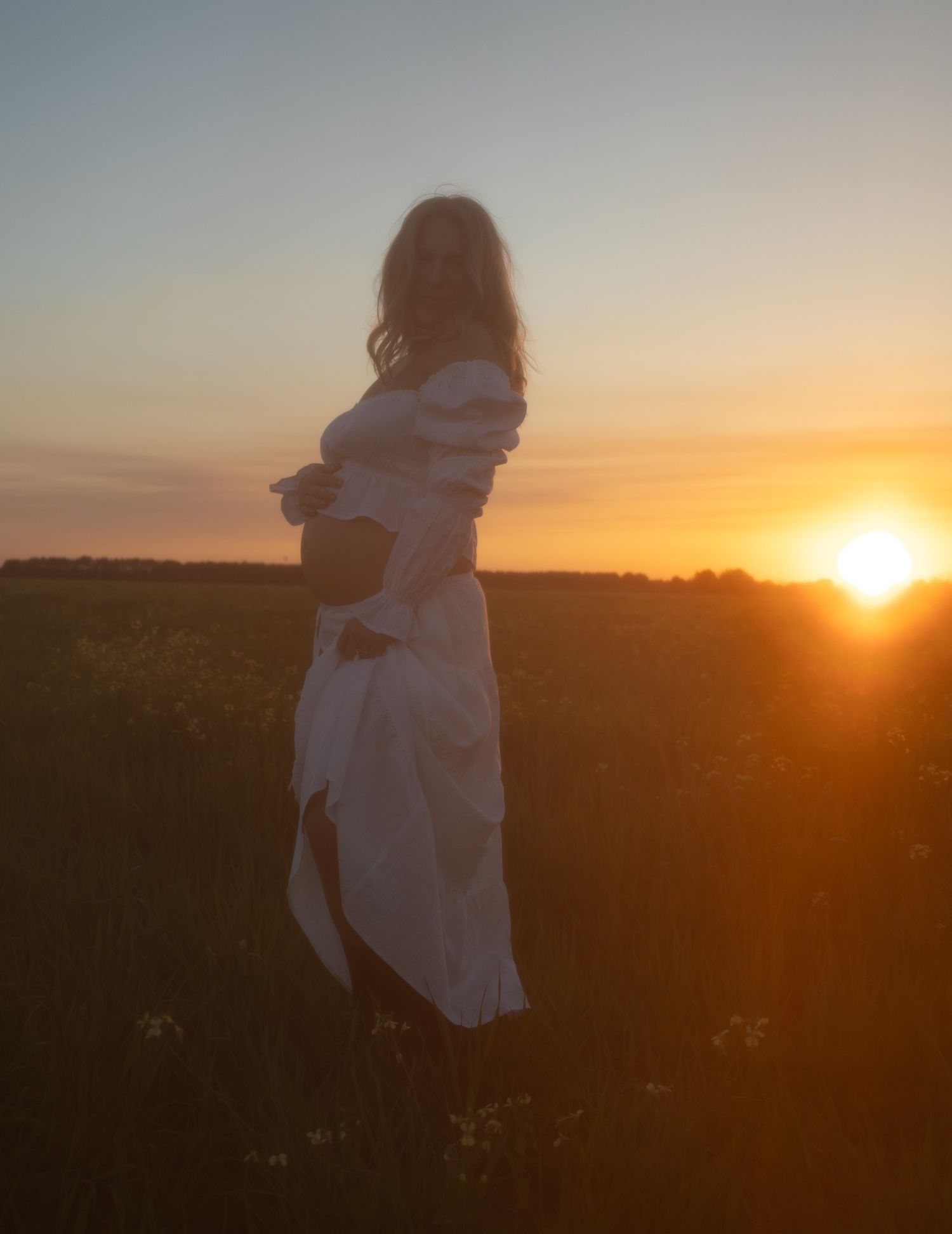 Pregnant woman in white dress silhouetted against dramatic golden sunset in open field.