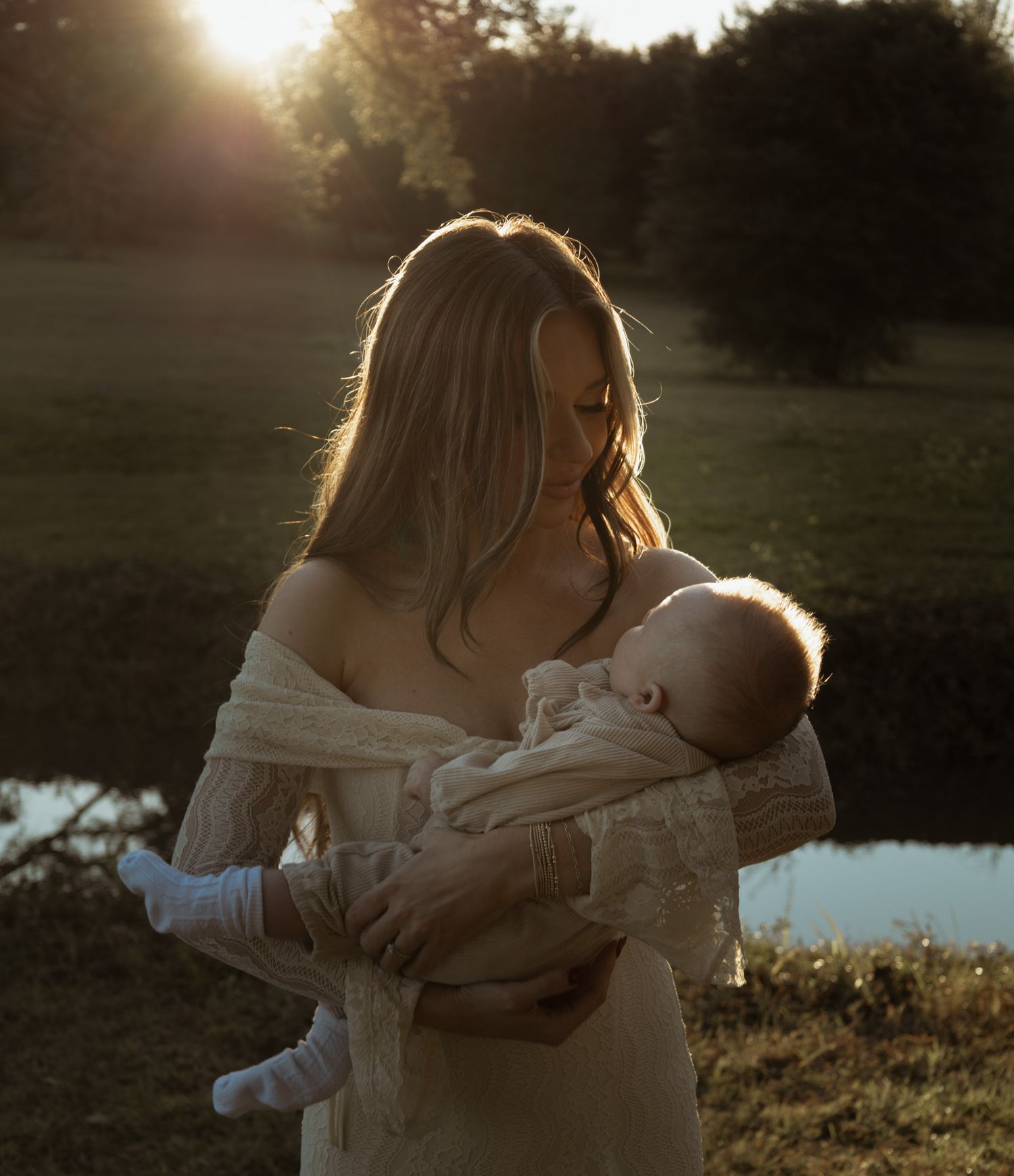 Young mother in off-shoulder dress tenderly holding sleeping newborn in golden hour light.