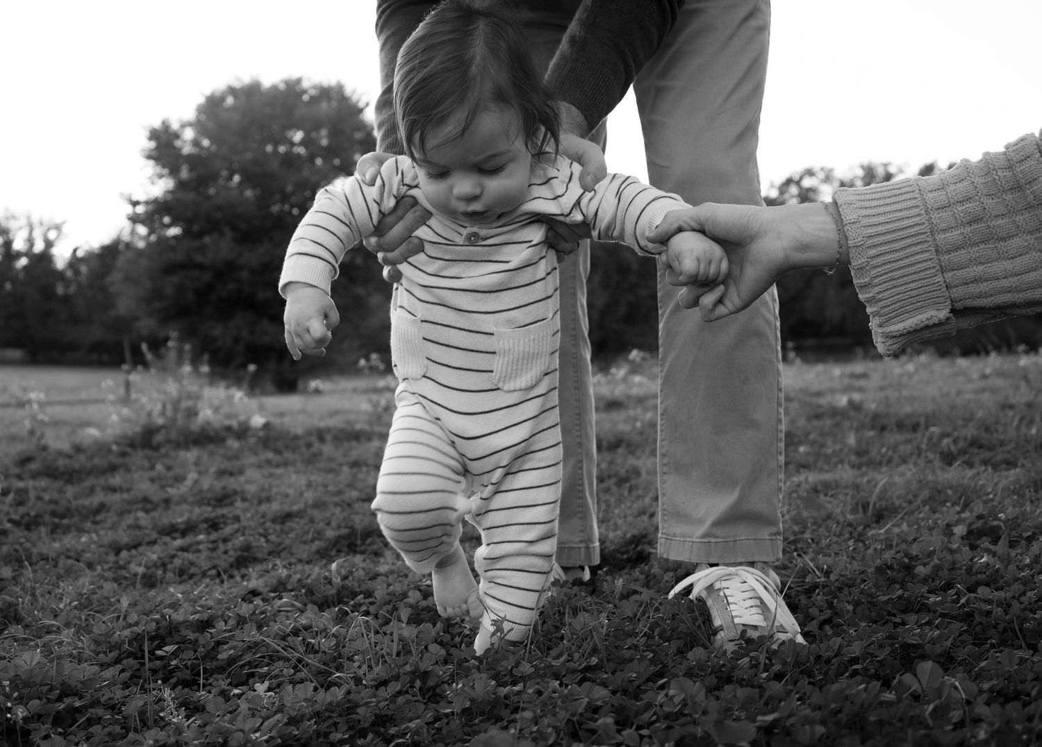A baby taking first steps on grass with parental support in a black and white photo.