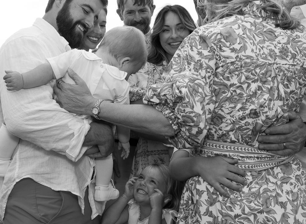 Candid black and white photo of joyful multigenerational family gathering around a baby outdoors.