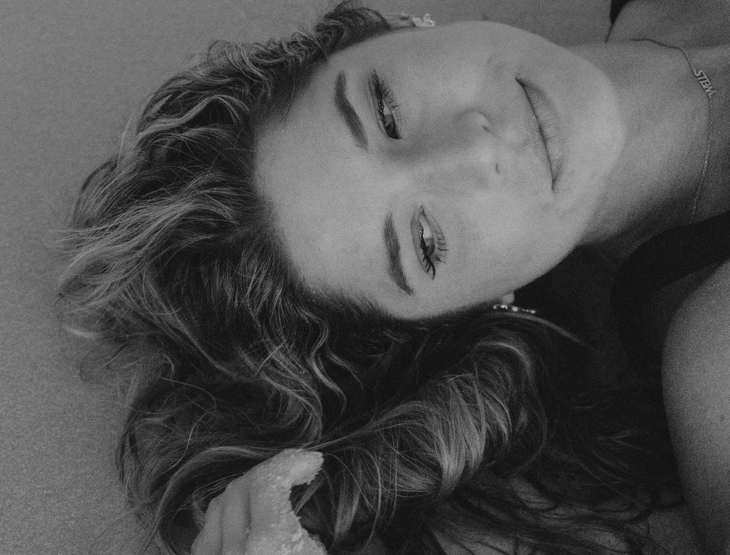 Close-up black and white portrait of a smiling woman lying on sand with wavy hair.