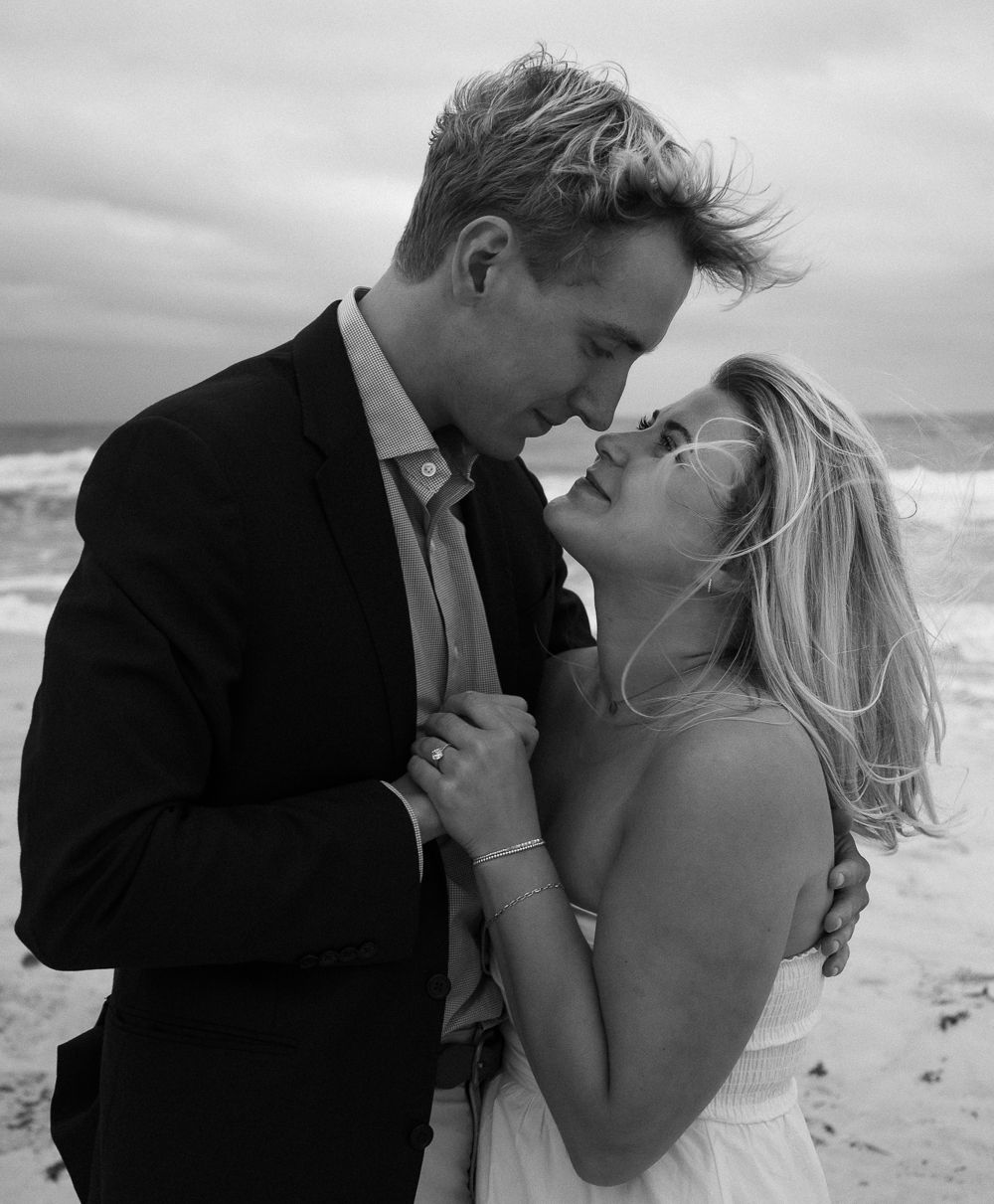 Black and white beach wedding photo of couple in formal attire sharing a romantic moment by the ocean waves.