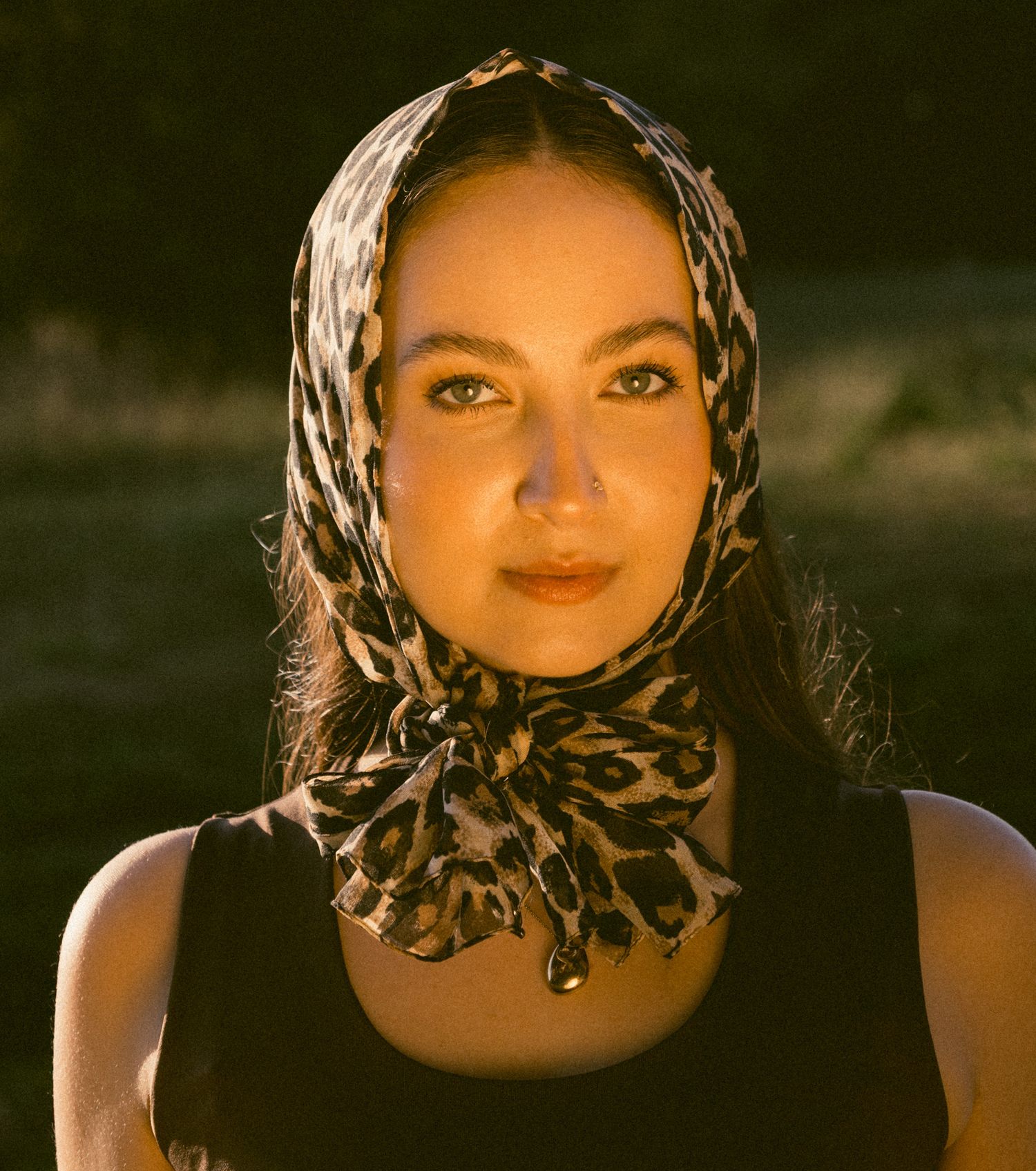Woman wearing leopard print headscarf and black tank top in warm golden hour light, front portrait.