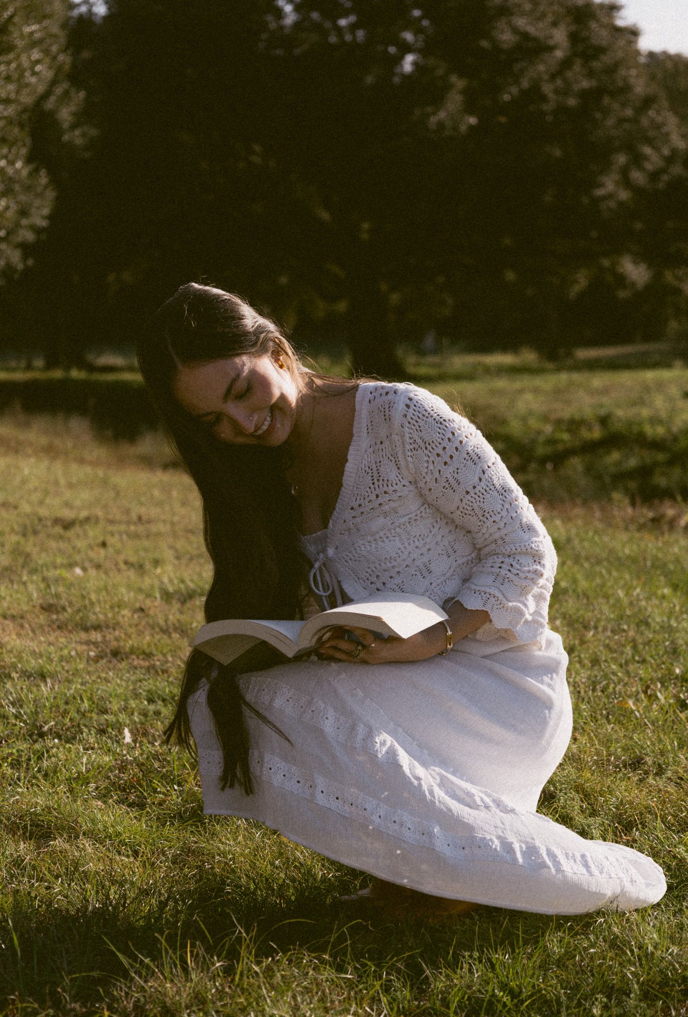 Young woman in white crochet dress smiling while reading a book outdoors on green grass.