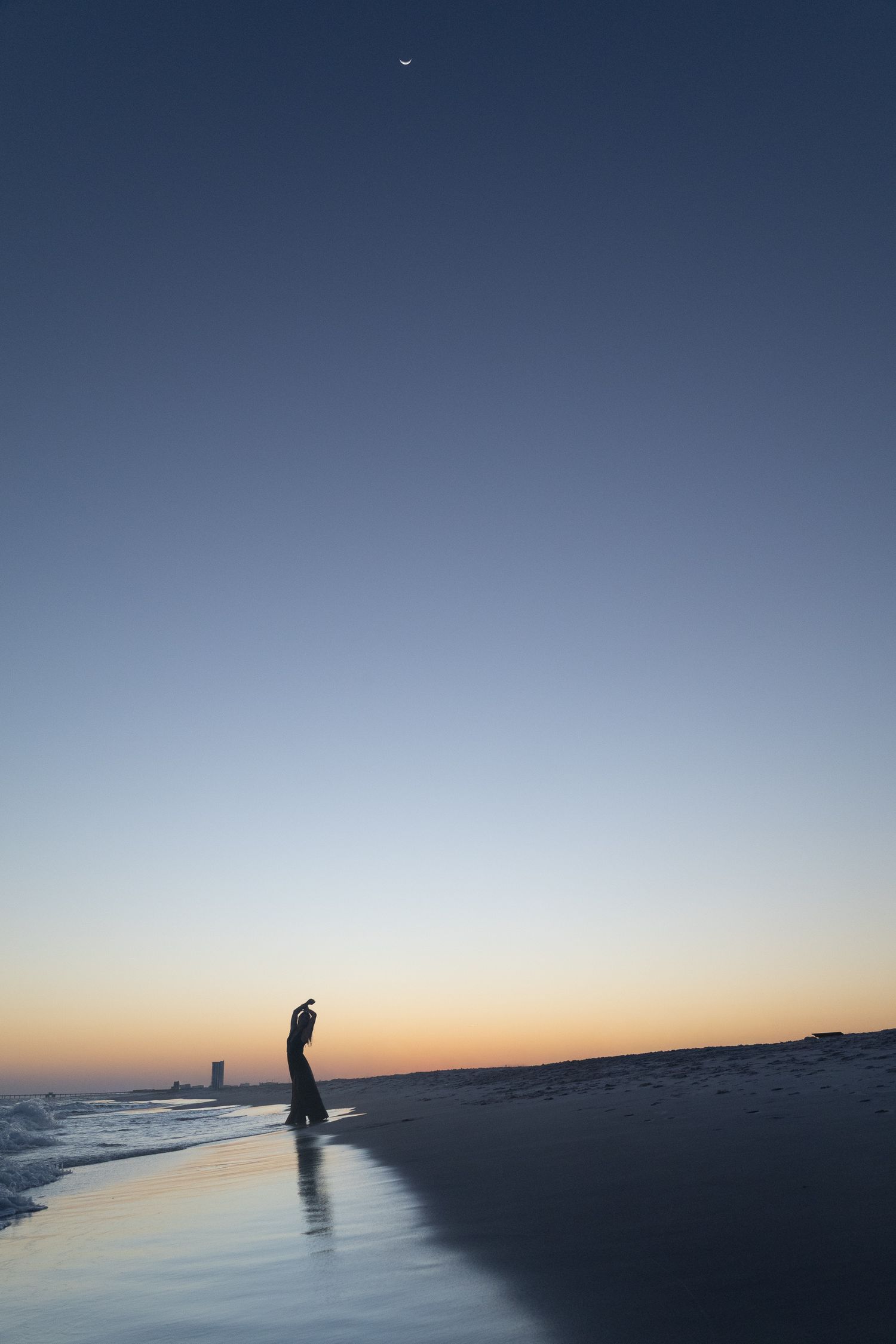 Silhouette of a person standing on beach at twilight with crescent moon in clear sky.