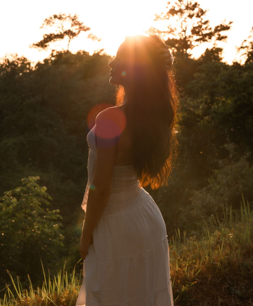 Woman in white dress standing in golden sunlight with sun flare filtering through forest trees.