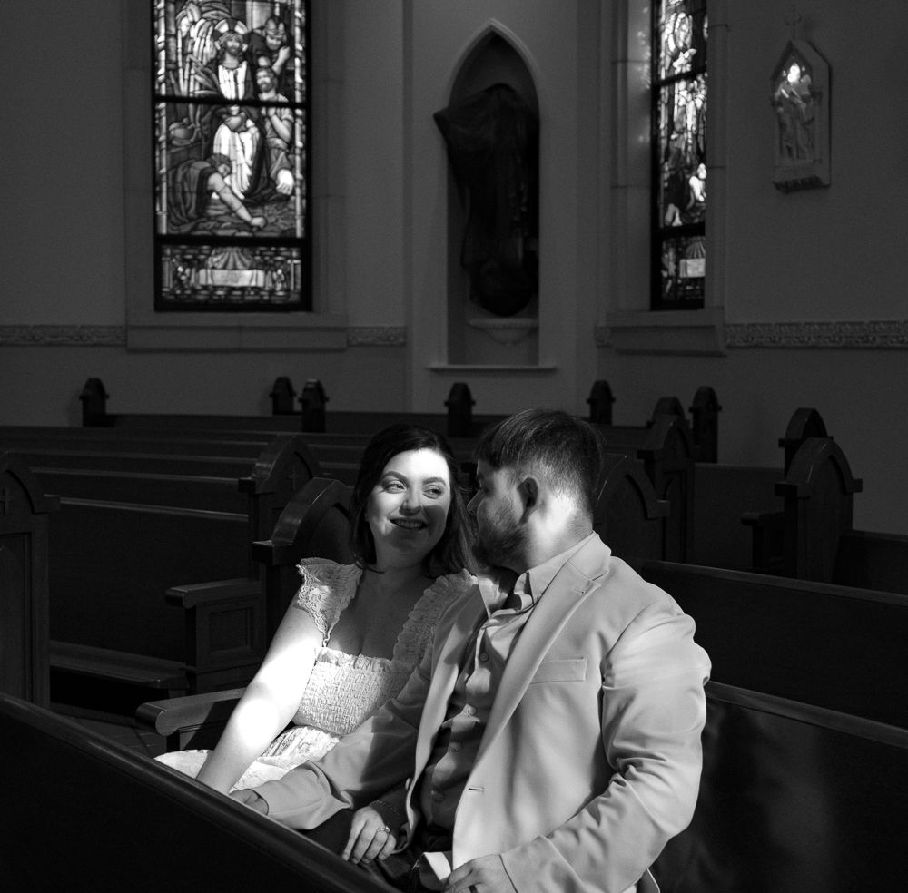 Black and white photo of a smiling bride and groom sitting together in a church pew.