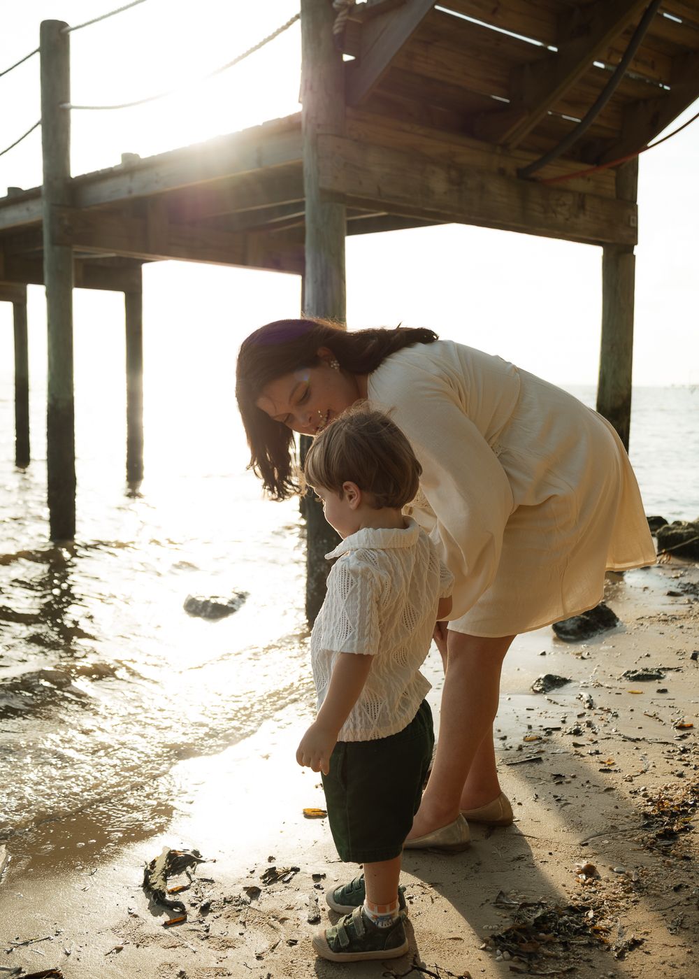A mother and toddler explore the sandy shore beneath a wooden pier at golden hour.