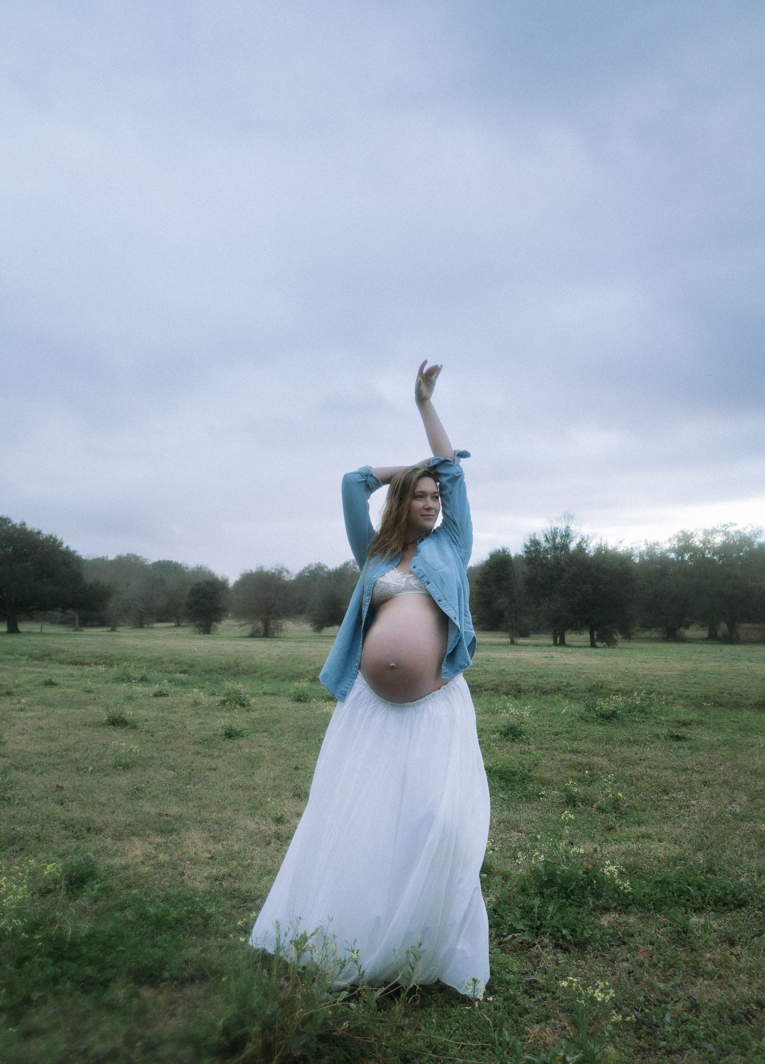 Pregnant woman in denim shirt and white skirt posing with arms raised in misty field.