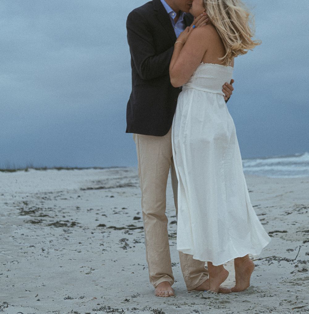 Couple embraces and kisses on a sandy beach at dusk, woman in flowing white dress barefoot.
