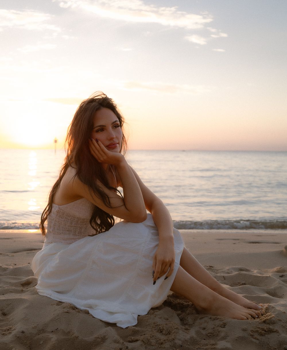 Beautiful brunette woman in white dress sitting on sandy beach during golden sunset by the ocean.