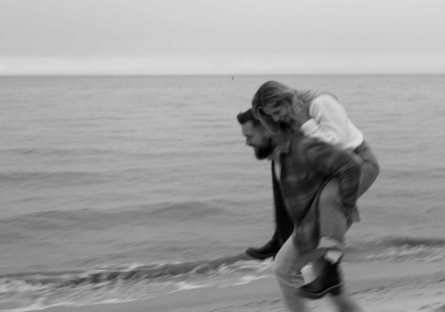 Black and white photo of a bearded man carrying a woman on his back while running along a beach shoreline.